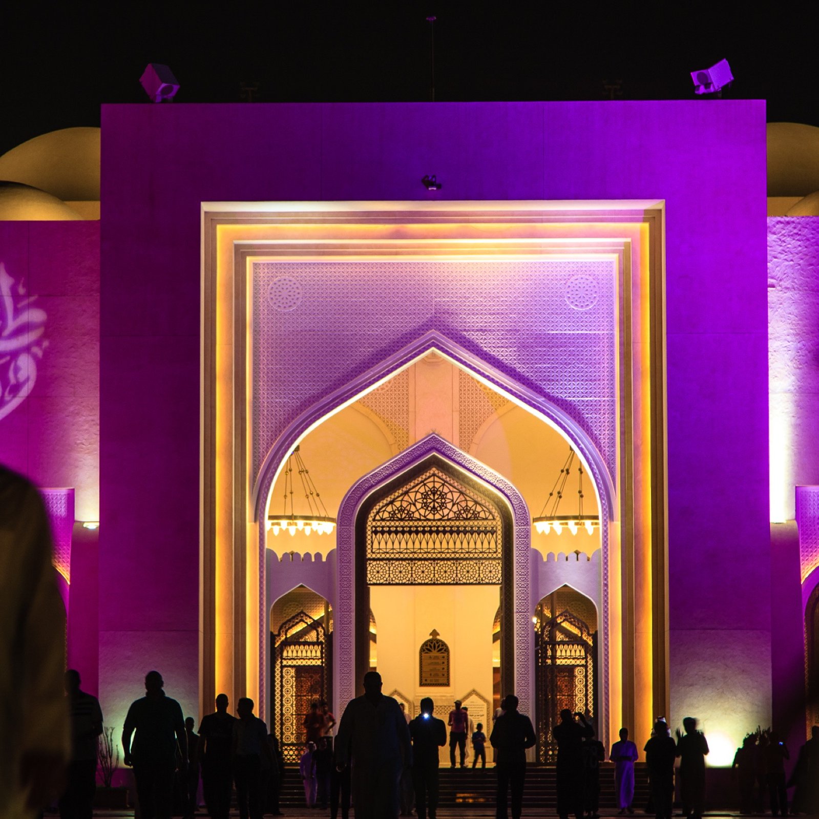 L'image montre une grande structure architecturale éclairée de manière colorée, principalement en violet. On peut voir un portail monumental avec des motifs décoratifs, typiques de l'architecture islamique. À l'avant, des silhouettes de personnes marchent, créant une atmosphère vivante et dynamique. Au fond, des éléments de design comme des arcs et des motifs sont visibles, ajoutant une touche d'élégance à l'ensemble de la scène. L'éclairage nocturne met en valeur les détails architecturaux et crée une ambiance fascinante.