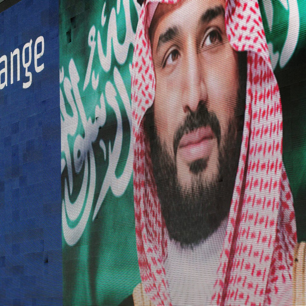 L'image montre une grande affiche murale présentant un homme en tenue traditionnelle saoudienne, avec un keffieh sur la tête. À côté de son visage, il y a un message en anglais qui dit "He is bringing change to Saudi Arabia" et un hashtag "#NewSaudiArabia". L'arrière-plan de l'affiche est bleu et le texte blanc. Cela évoque un thème de changement et de modernisation en Arabie Saoudite.