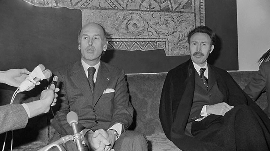 L'image montre deux hommes assis sur un canapé, apparemment lors d'une conférence de presse ou d'une rencontre. Ils semblent sérieux et attentifs, avec des microphones en face d'eux, indiquant qu'ils sont en train de répondre à des questions. L'un des hommes porte un costume sombre et a une expression réfléchie, tandis que l'autre, à sa droite, est vêtu d'une façon plus formelle, peut-être avec une écharpe ou un manteau. En arrière-plan, il y a un motif mural qui ajoute une certaine ambiance à la scène.