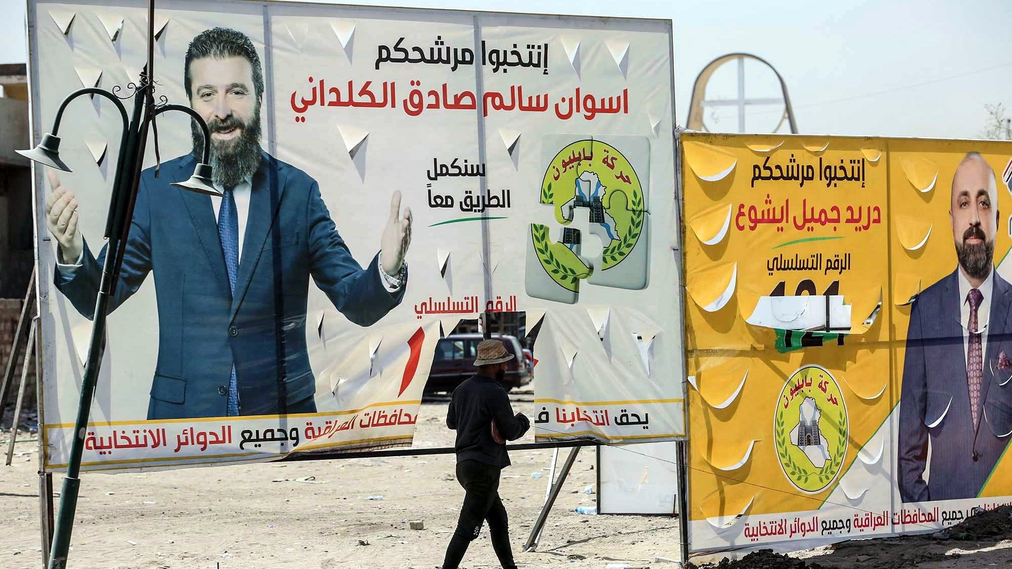 تظهر الصورة شخصًا يسير بجانب لوحات إعلانات كبيرة تتضمن صورًا وعناوين تتعلق بانتخابات. إحدى اللوحات تحتوي على صورة لرجل يُدعى سالم صادق الكلداني، والأخرى تتعلق بشخص يُدعى جليل بشير. النص في الإعلانات يتضمن عبارات تدعو الناخبين لاختيار المرشحين للمزيد من التفاعل والمشاركة السياسية. الخلفية تسلط الضوء على أجواء انتخابية تشهدها منطقة معينة.