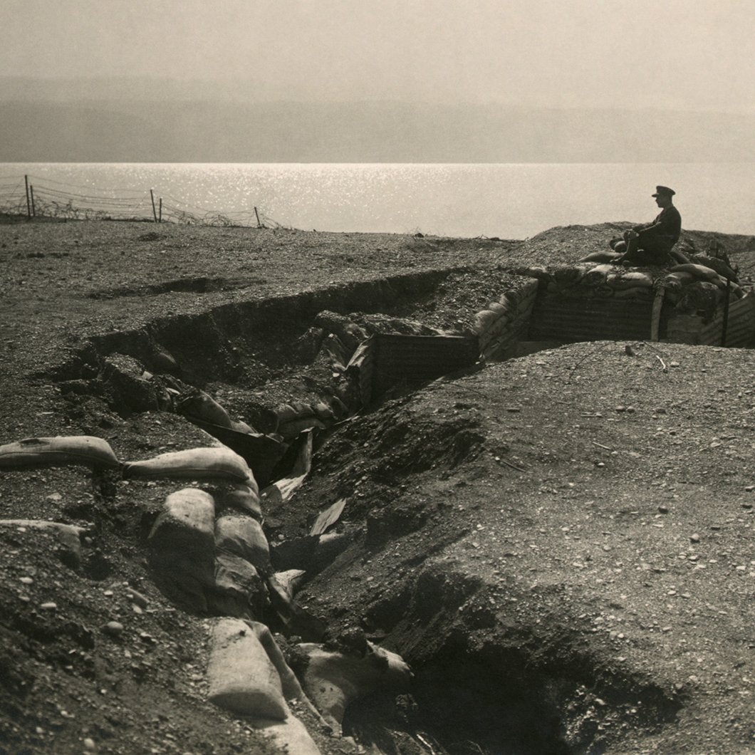 L'image représente un champ de bataille avec une tranchée. On peut voir un soldat assis sur les bords de la tranchée, qui semble surveiller les environs. Le terrain est accidenté et semé de sacs de sable, typiques des fortifications de la Première Guerre mondiale. À l'arrière-plan, on aperçoit une étendue d'eau qui pourrait être un lac ou une rivière. L'atmosphère générale est sombre et mélancolique, évoquant la dureté de la guerre.