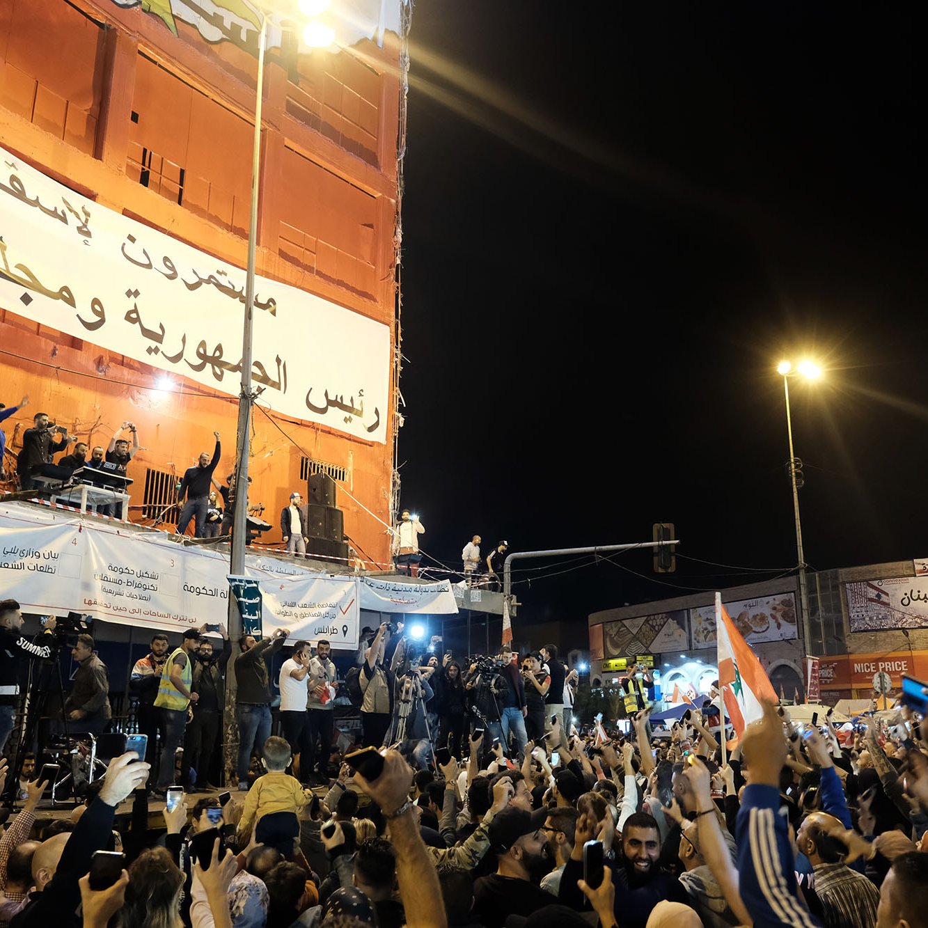 The image captures a large crowd gathered in an urban area, likely during a rally or protest. People are raising their hands, some holding flags, and others are holding up their phones, possibly recording the event. In the background, there is a large banner that appears to convey a political message, and a group of speakers is visible on a stage or elevated platform. The atmosphere seems energetic and filled with a sense of purpose, with bright lights illuminating the scene at night.