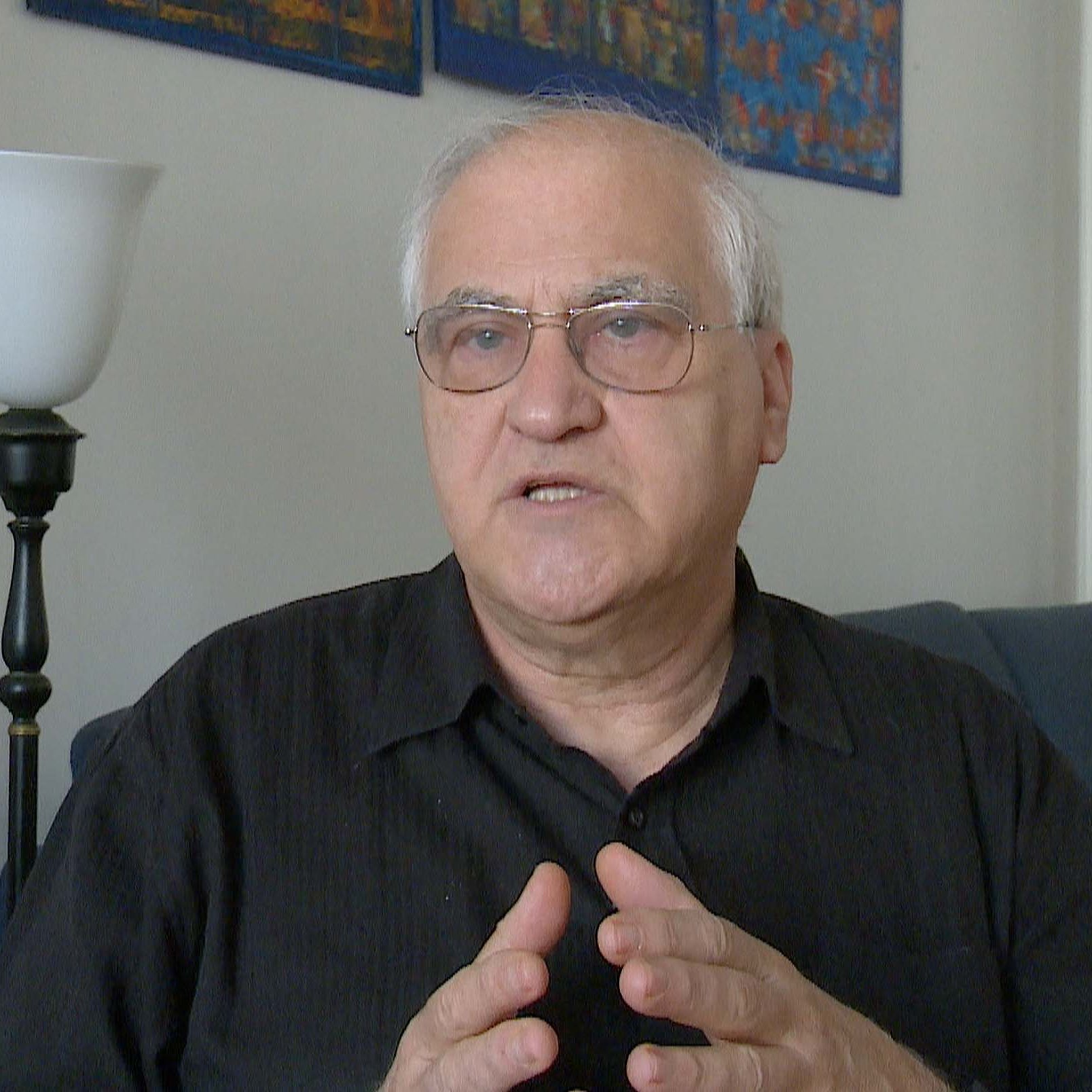 L'image montre un homme parlant avec des mains gesticulant, exprimant probablement une idée ou un point de vue. Il porte des lunettes et une chemise noire, et il semble attentif à la conversation. En arrière-plan, il y a un canapé bleu et une source de lumière, ce qui indique un environnement intérieur lumineux et accueillant. Des œuvres d'art sont accrochées au mur, ajoutant une touche décorative à la scène.