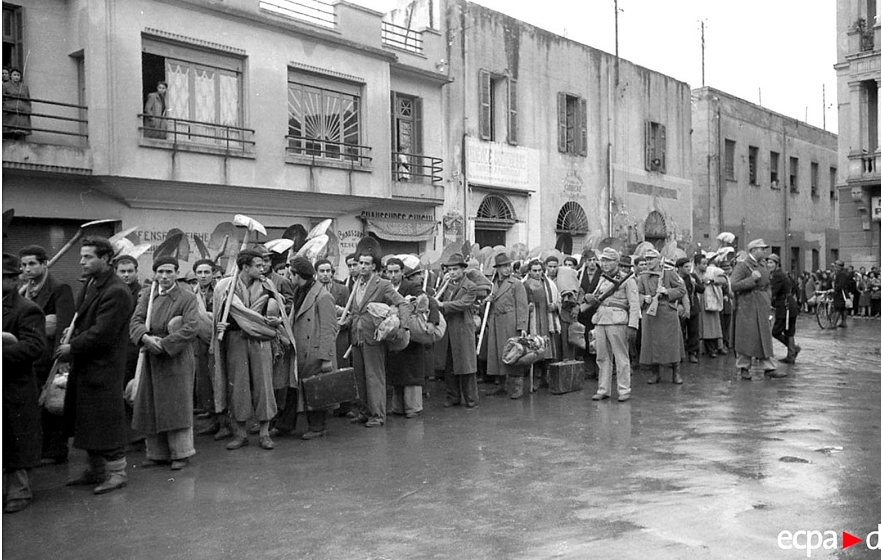 L'image montre une file de personnes alignées dans une rue apparemment urbaine. Les individus portent des vêtements de travail et des sacs, ce qui suggère qu'ils attendent quelque chose, peut-être un transport ou un appel. En arrière-plan, on peut voir des bâtiments avec des fenêtres et des balcons, ainsi que des éléments architecturaux qui indiquent une époque passée. L'atmosphère semble sérieuse, et le pavement est mouillé, peut-être à cause de la pluie.