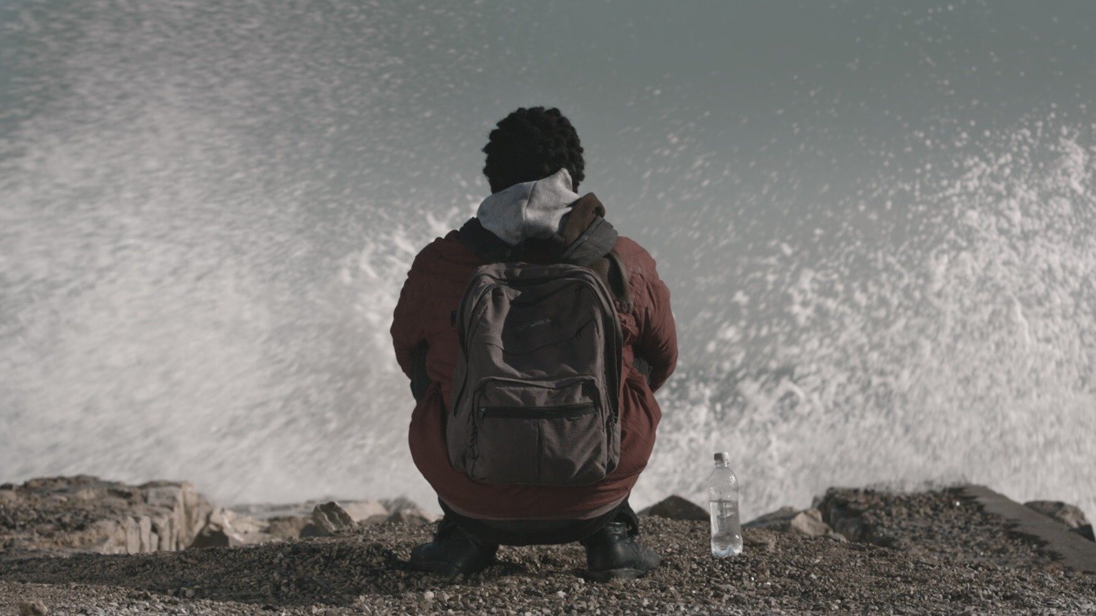 في الصورة، يظهر شخص يجلس على حافة شاطئ أو منطقة قريبة من البحر. يبدو أنه شاب ذو شعر مجعد، ويرتدي معطفًا سميكًا مع كيان داخلي ورأسه مائل قليلاً إلى الأمام. خلفه، تتلاطم أمواج البحر بقوة، مما يعطي إحساسًا بالحركة والطبيعة القوية. بجانبه، يوجد زجاجة ماء على الأرض. الأجواء تبدو هادئة بالرغم من قوة الأمواج، مما يضفي شعورًا بالتأمل أو الوحدة.