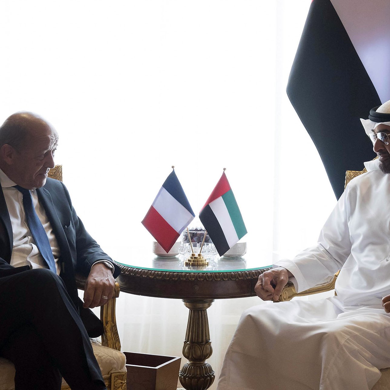 The image shows two men seated at a table in a luxurious setting. They are engaged in conversation, appearing to be in a diplomatic or formal meeting. There are flags on the table representing France and the United Arab Emirates. The background features elegant curtains and decor. One man is dressed in a suit, while the other is wearing traditional Emirati attire.