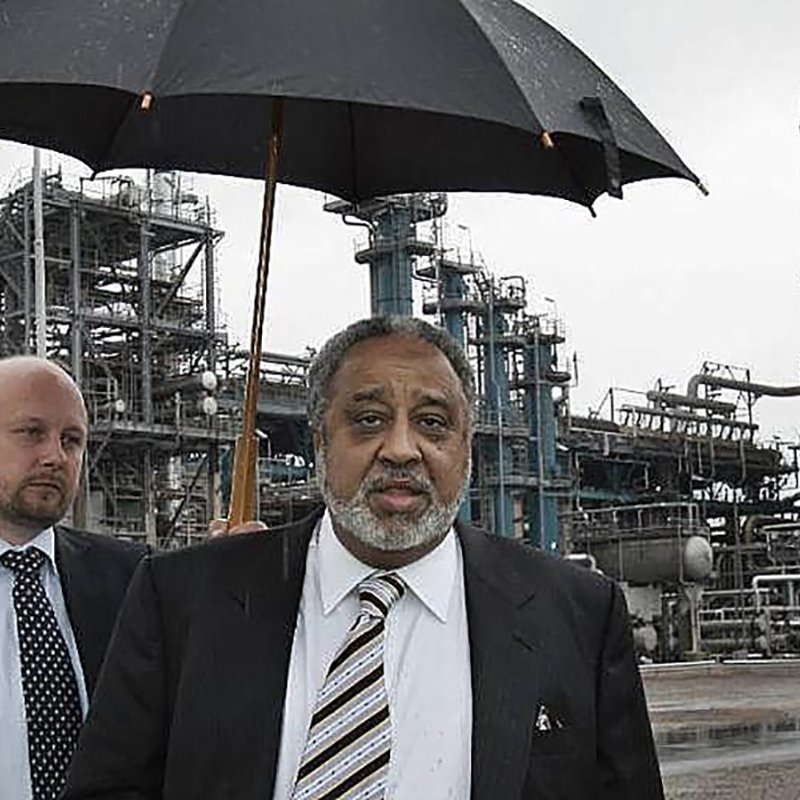 L'image montre deux hommes marchant dans une zone industrielle. L'un des hommes, qui porte un costume noir et une cravate, tient un parapluie, probablement pour se protéger de la pluie. L'autre homme, à l'arrière-plan, est également en costume. On peut voir des structures industrielles, comme des réservoirs et des cheminées, en arrière-plan, sous un ciel grisâtre. L'ambiance générale de l'image est celle d'une visite ou d'une inspection dans un environnement industriel.