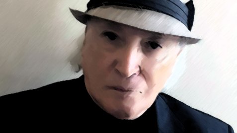 الصورة تظهر شخصًا يرتدي قبعة رمادية مع شريط داكن، وهو يرتدي سترة داكنة أيضًا. لديه ملامح وجه جادة، والخلفية تبدو بسيطة وغير مزدحمة، مما يركز الانتباه على تعبيره وملابسه. توزيع الإضاءة في الصورة يبرز ملامحه بشكل واضح.