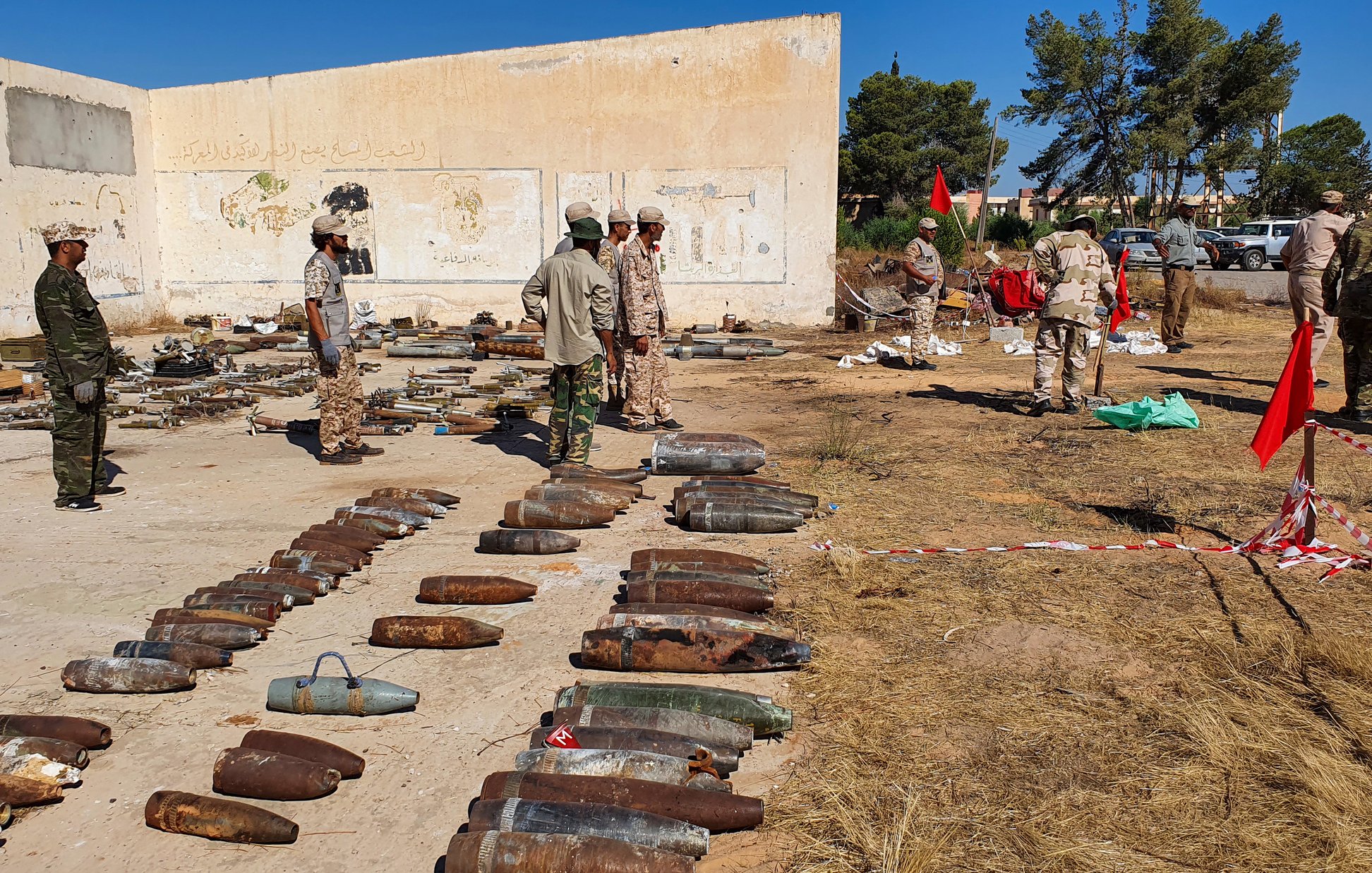 L'image montre un site en extérieur où des bombes et des munitions sont disposées au sol. Plusieurs personnes en uniformes militaires sont présentes, certaines examinant les objets, tandis que d'autres semblent discuter ou travailler sur le terrain. En arrière-plan, on peut voir un mur partiellement recouvert de graffitis et des arbres qui entourent la zone. Des drapeaux sont également visibles, et le sol est marqué par des restes de munitions, signifiant probablement un site de déminage ou de collecte de matériel explosif.