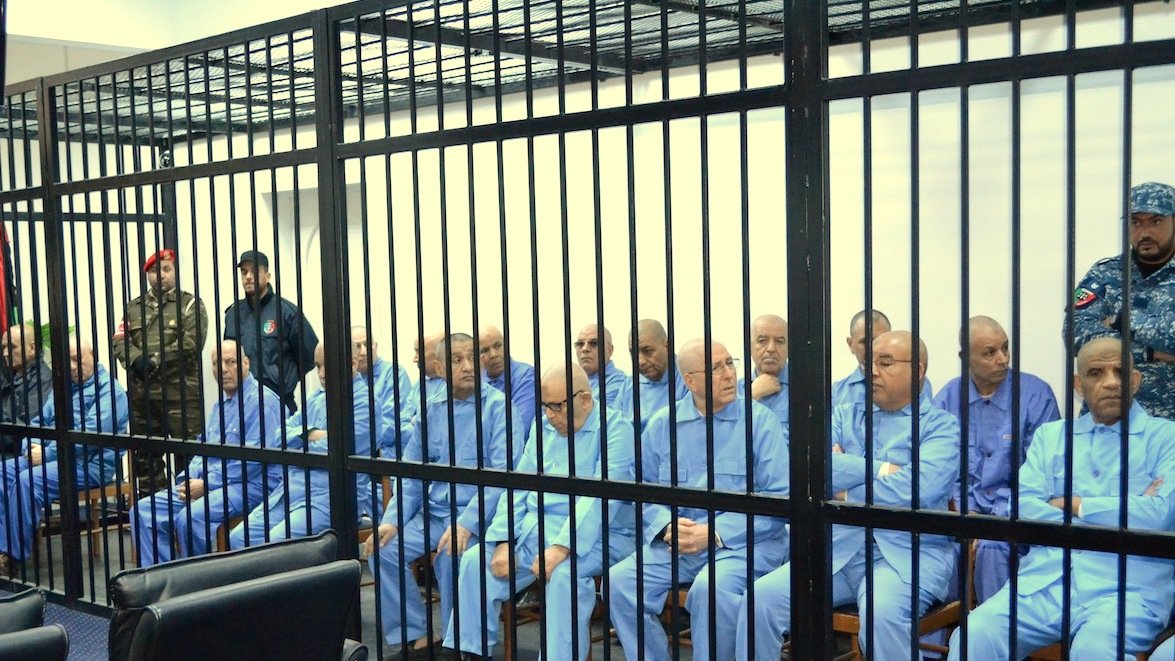 L'image montre un groupe d'individus assis derrière des barreaux. Ils portent tous des vêtements similaires, de couleur bleue. L'atmosphère semble sérieuse et formelle, peut-être en lien avec une audience ou un procès. Plusieurs personnes en uniforme sont présentes, suggérant un contexte de sécurité ou d'autorité. Les individus semblent concentrés ou préoccupés.