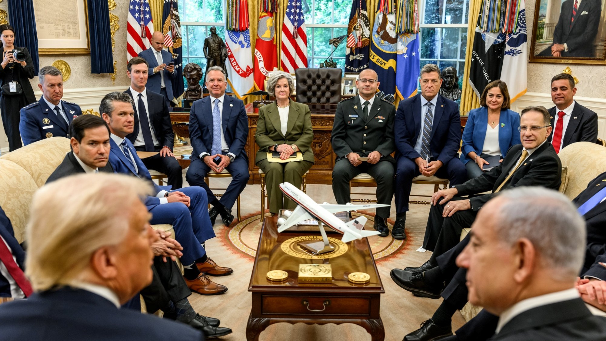 A formal meeting in the Oval Office with various officials and a model airplane on the table.