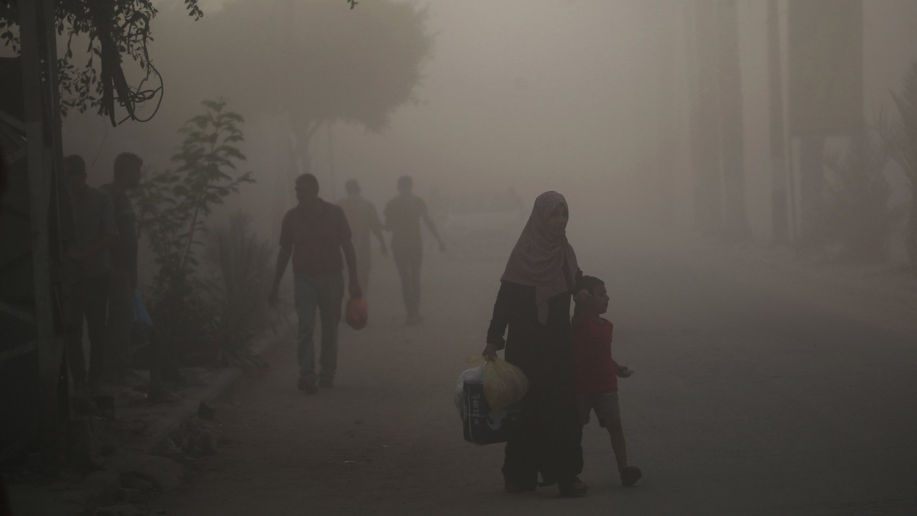 L'image montre une scène urbaine plongée dans une atmosphère brumeuse ou poussiéreuse. Dans le premier plan, une femme portant un hijab tient la main d'un jeune garçon. Ils semblent marcher ensemble le long de la rue. En arrière-plan, d'autres silhouettes de personnes sont visibles, floues par la brume, tandis que des arbres et des structures urbaines apparaissent à peine à travers la poussière. L'ambiance semble calme mais chargée d'un certain mystère à cause de la visibilité réduite.