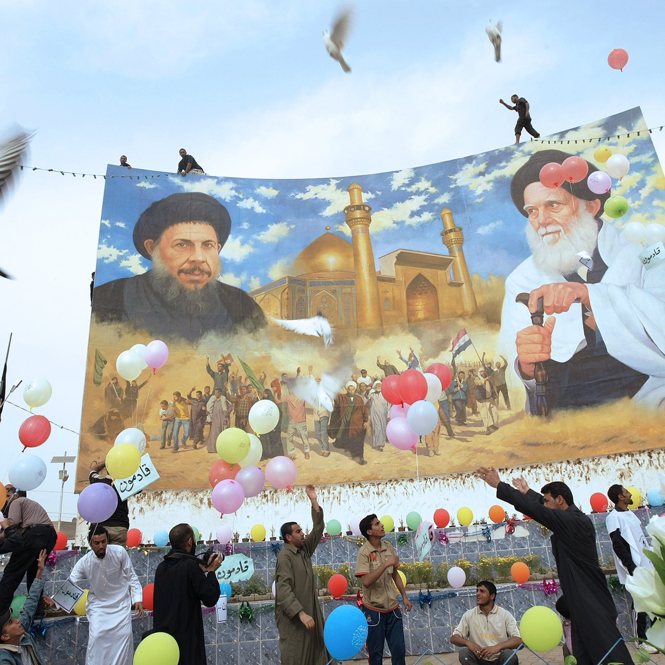 تظهر الصورة احتفالًا جماهيريًا حيث يحتشد مجموعة من الأشخاص تحت لافتة كبيرة تحتوي على صور لشخصيتين بارزتين. يبدو أنهم يقومون بإطلاق بالونات ملونة، ومن حولهم أجواء من الفرح والاحتفالات. في الخلفية، يُمكن رؤية معلم معماري يُحتمل أن يكون مسجدًا أو مكانًا مقدسًا. الألوان الساطعة والأجواء الحيوية تعكس روح الاحتفال والوحدة في المجتمع.