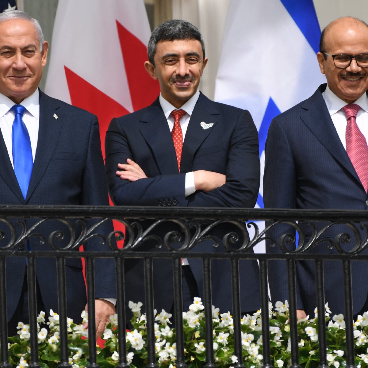 در این تصویر سه نفر در یک بالکن ایستاده‌اند. سمت چپ، مردی با کت و شلواری تیره و کراوات آبی قرار دارد. در وسط، مردی با لباس رسمی و کراوات قرمز دیده می‌شود که لبخند می‌زند. سمت راست، مردی دیگر در لباس رسمی با کراوات قرمز تیره ایستاده است. در پس‌زمینه، پرچم‌های کشورهای مختلف شامل ایالات متحده، کانادا و دیگر کشورها نمایان است. گل‌های سفیدی در نرده بالکن قرار دارند.