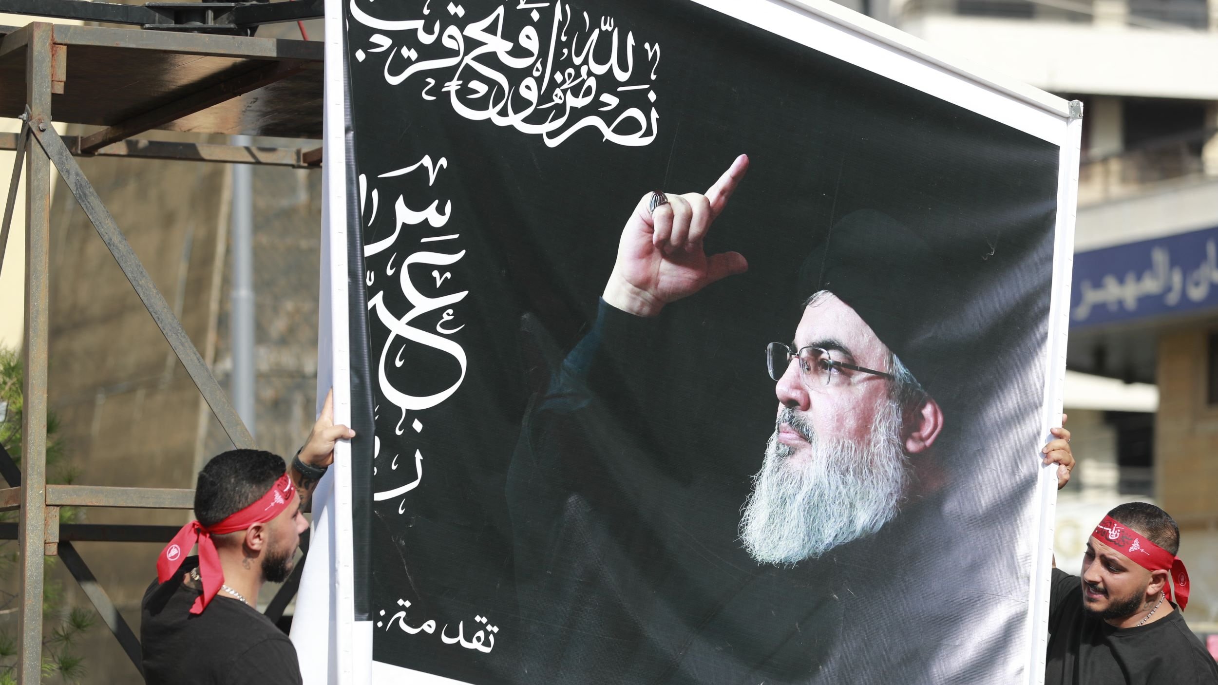 تصویر دو نفر را نشان می‌دهد که یک تابلو بزرگ را با تصویری از فردی در حال اشاره کردن به سمت بالا در دست دارند. پس‌زمینه تابلو سیاه است و بر روی آن متن‌هایی به زبان عربی دیده می‌شود. افراد حاضر در تصویر لباس‌های سیاه دارند و برخی از آن‌ها دستبندهای همرنگ بر روی سرشان دارند. این صحنه به نظر می‌رسد که در یک تجمع یا مراسم خاصی در حال برگزاری است.