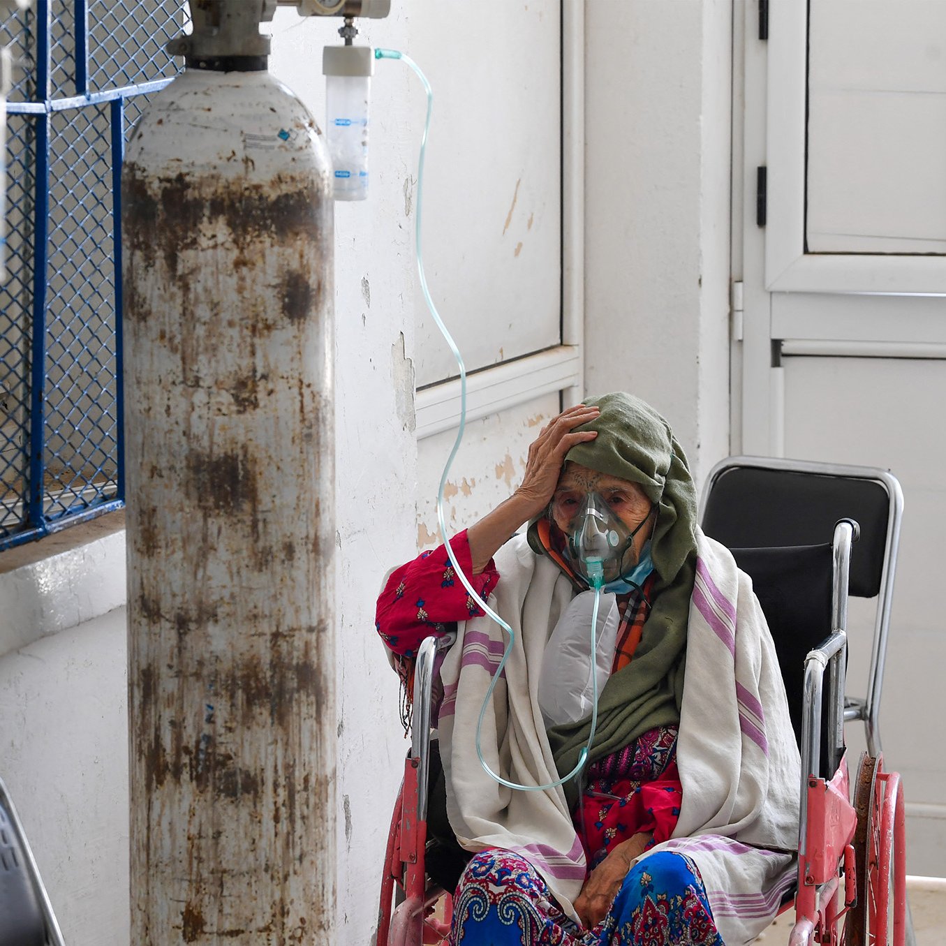 L'image montre une femme âgée assise dans un couloir d'hôpital. Elle porte un masque et une écharpe sur la tête. On peut voir des bouteilles d'oxygène à proximité, indiquant qu'elle reçoit des soins médicaux. L'environnement est simple, avec des murs blancs et des fenêtres qui laissent passer la lumière. L'expression de la femme suggère une certaine inquiétude ou fatigue.