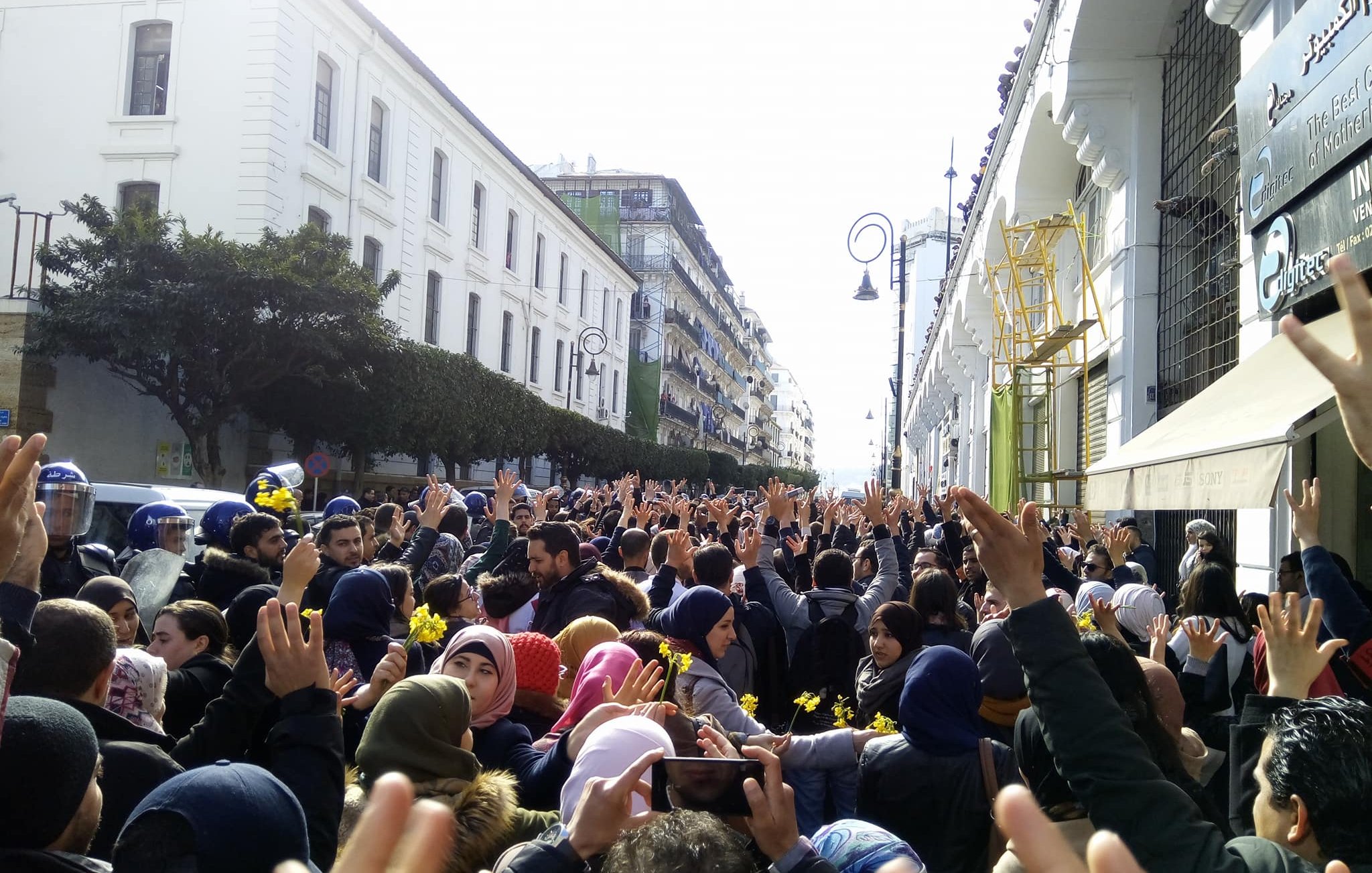 The image depicts a large crowd of people gathered in an urban setting, possibly during a protest or demonstration. Many individuals are raising their hands, and a sense of unity and collective action is present. The atmosphere appears to be lively, and the crowd is surrounded by buildings, suggesting a city environment. Some police presence is visible, indicating that the gathering may be significant or organized and possibly involves a call for change or social issues. The crowd includes diverse individuals, wearing a range of clothing styles, with some wearing headscarves.