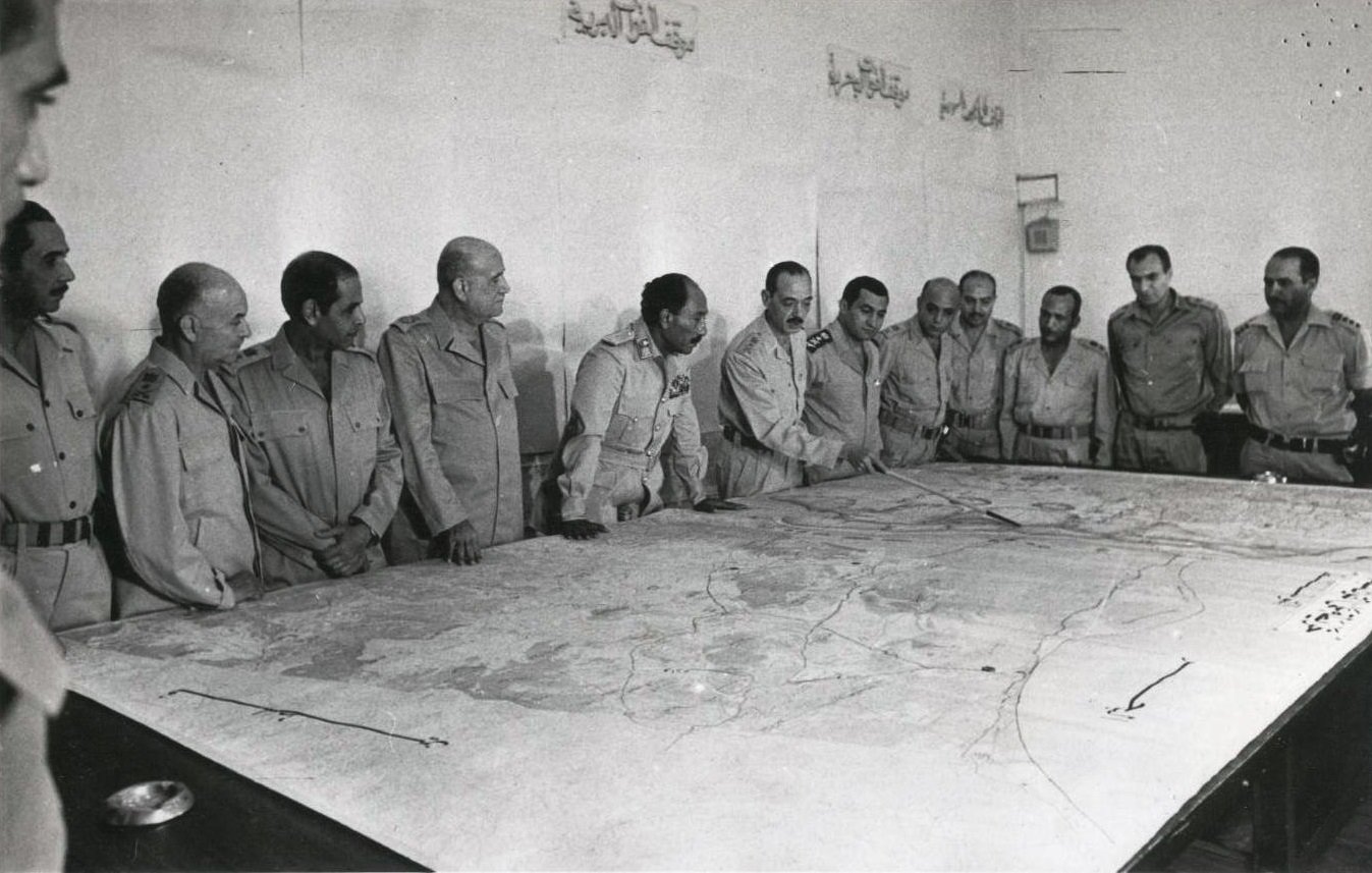 تظهر الصورة مجموعة من الرجال يرتدون زيًا عسكريًا، يجتمعون حول طاولة كبيرة تعرض خريطة. يبدو أن الاجتماع يدور حول التخطيط أو مناقشة استراتيجية ما. تعكس تعابير وجوههم التركيز والاهتمام بما يُناقش. الجدران خلفهم تبدو بسيطة، مع بعض الكتابات باللغة العربية. تُعطي الصورة انطباعًا عن موقف رسمي وعسكري.