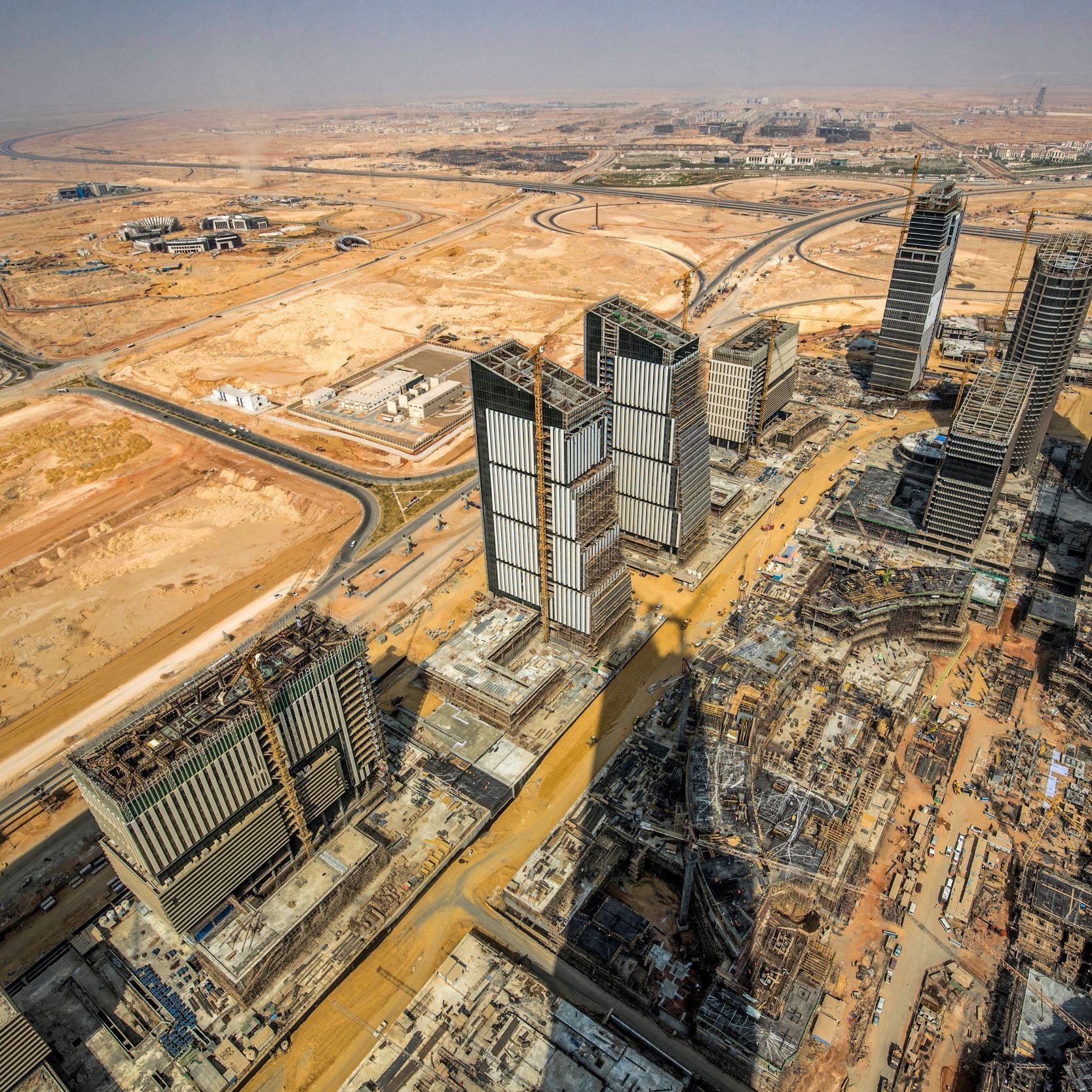 The image showcases a panoramic view of a vast construction site in a desert landscape. In the foreground, several high-rise buildings are partially completed, with cranes and construction equipment evident. The surrounding area features empty land and some roadways, characteristic of a developing urban environment. The backdrop includes a clear sky and distant structures, suggesting a mixture of ongoing urbanization and natural desert terrain. The overall scene illustrates the rapid expansion of urban infrastructure in a typically arid region.