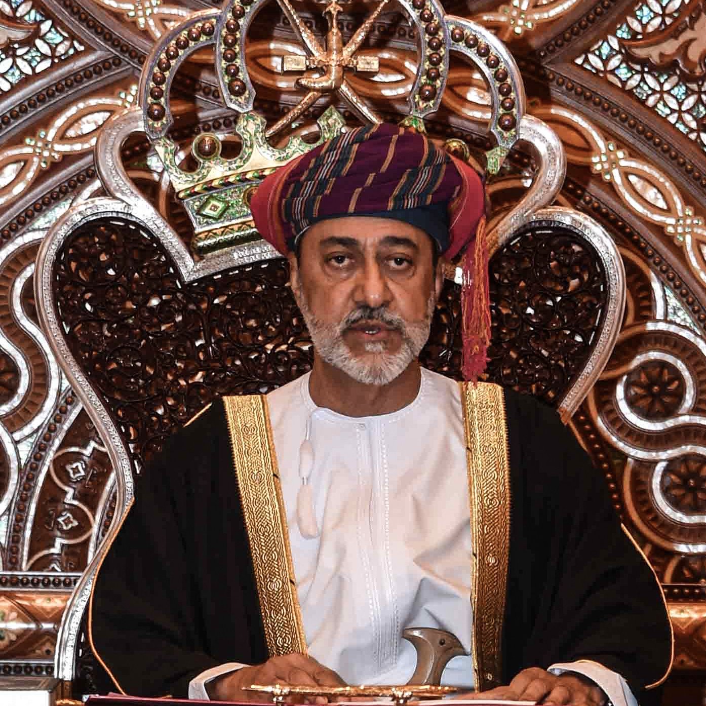 L'image montre un homme assis sur un trône orné. Il porte un costume traditionnel, comprenant une robe blanche et un turban coloré. Son expression est sérieuse et solennelle. Le fond est richement décoré avec des motifs complexes, créant une ambiance majestueuse. Les détails de la couronne et du trône mettent en valeur son statut élevé.