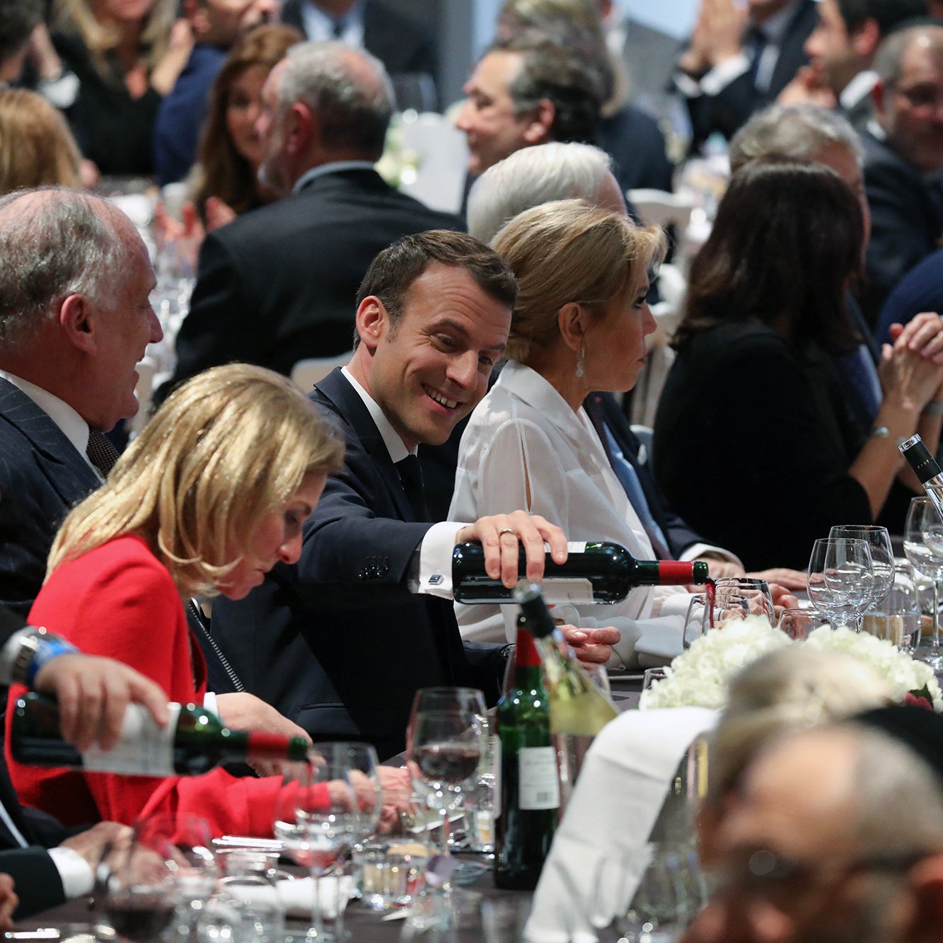L'image montre une scène de dîner élégant, probablement lors d'un événement officiel. Plusieurs personnes sont assises autour d'une grande table, décorée avec des verres, des bouteilles de vin et des fleurs. Au centre, on aperçoit un homme souriant en train de servir du vin, semblant profiter de l'atmosphère. Les convives sont habillés de manière formelle, et l'ambiance paraît conviviale et animée, avec des conversations visibles parmi les invités.