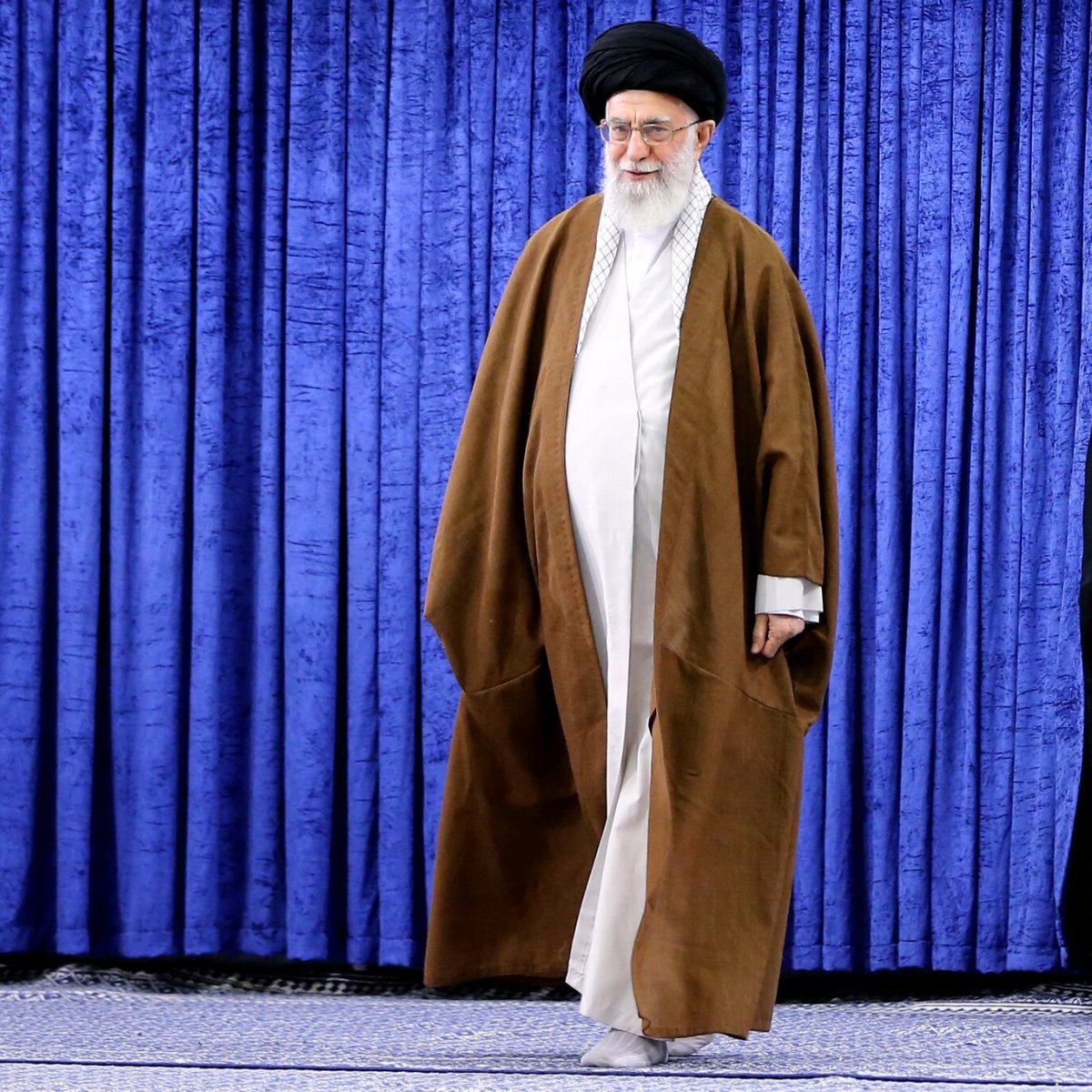 در تصویر، فردی با لباس سنتی روحانی به رنگ‌های قهوه‌ای و سفید دیده می‌شود. او کلاهی مشکی بر سر دارد و ریش بلندی دارد. پس‌زمینه تصویر شامل پرده‌ای به رنگ آبی است. فرد در حال حرکت به سمت سمت راست تصویر قرار دارد. این تصویر به طور کلی حس وقار و احترام را القا می‌کند.