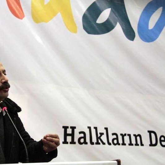 L'image montre un homme parlant lors d'un événement ou d'une conférence. Il est vêtu d'un manteau sombre et d'une chemise blanche. Le fond affiche une grande bannière avec des mots colorés, dont "Halkların Derneği", qui signifie "L'Association des Peuples" en turc. L'homme semble engagé dans son discours, utilisant des gestes pour illustrer ses propos.