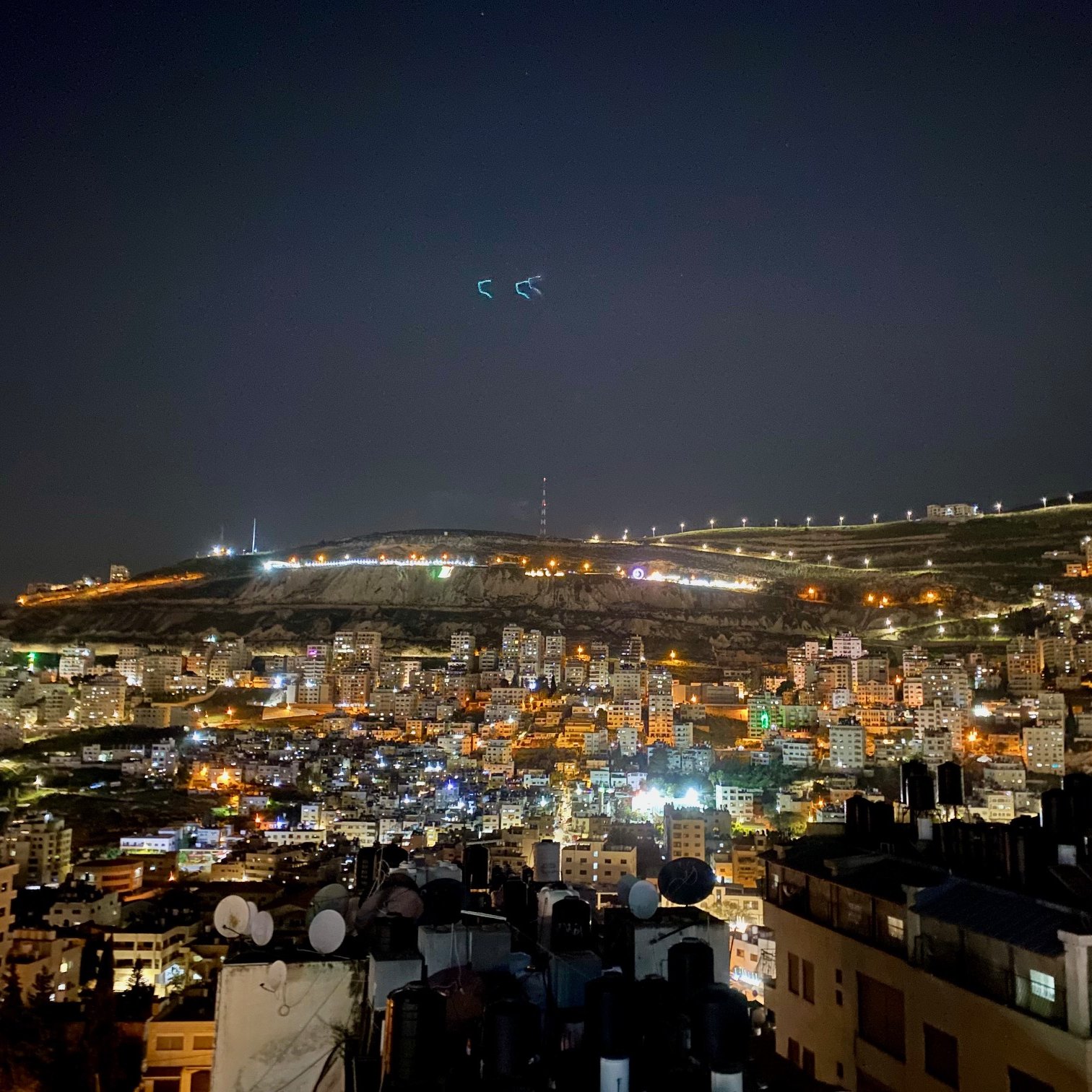 The image shows a nighttime view of a cityscape, likely on the slope of a hill or mountain. The scene is illuminated with numerous lights from buildings, giving it a vibrant and lively atmosphere. In the foreground, there are rooftops with antennas or satellite dishes visible, while the background features the illuminated hillside, possibly with telecommunications towers or other structures visible at the top. The sky is dark, suggesting it's a clear night with some stars or light reflections. Overall, the image conveys a sense of urban life and a stunning nocturnal landscape.