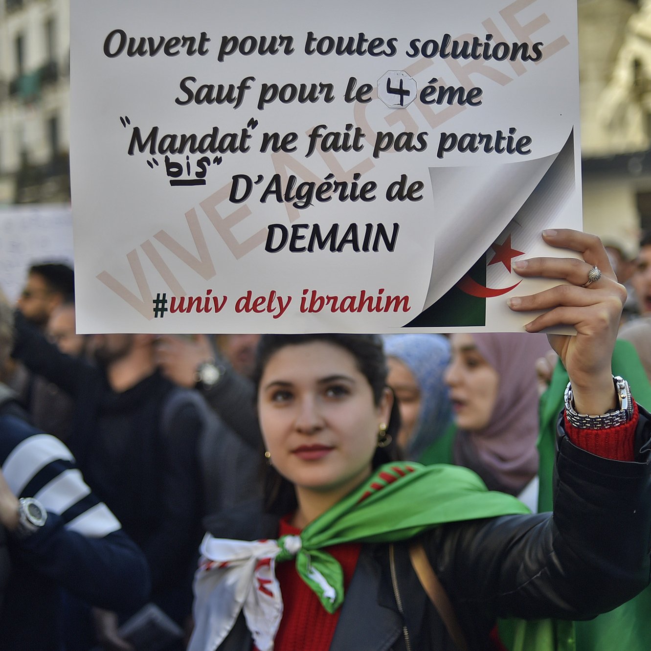 L'image montre une manifestation, où une jeune femme tient une pancarte. La pancarte indique qu'elle est ouverte à toutes solutions, sauf pour le 4ème mandat, affirmant que le mandat ne fait pas partie de l'Algérie de demain. La femme est entourée d'autres manifestants et porte un drapeau. L'atmosphère semble engagée et déterminée, reflétant des revendications politiques.