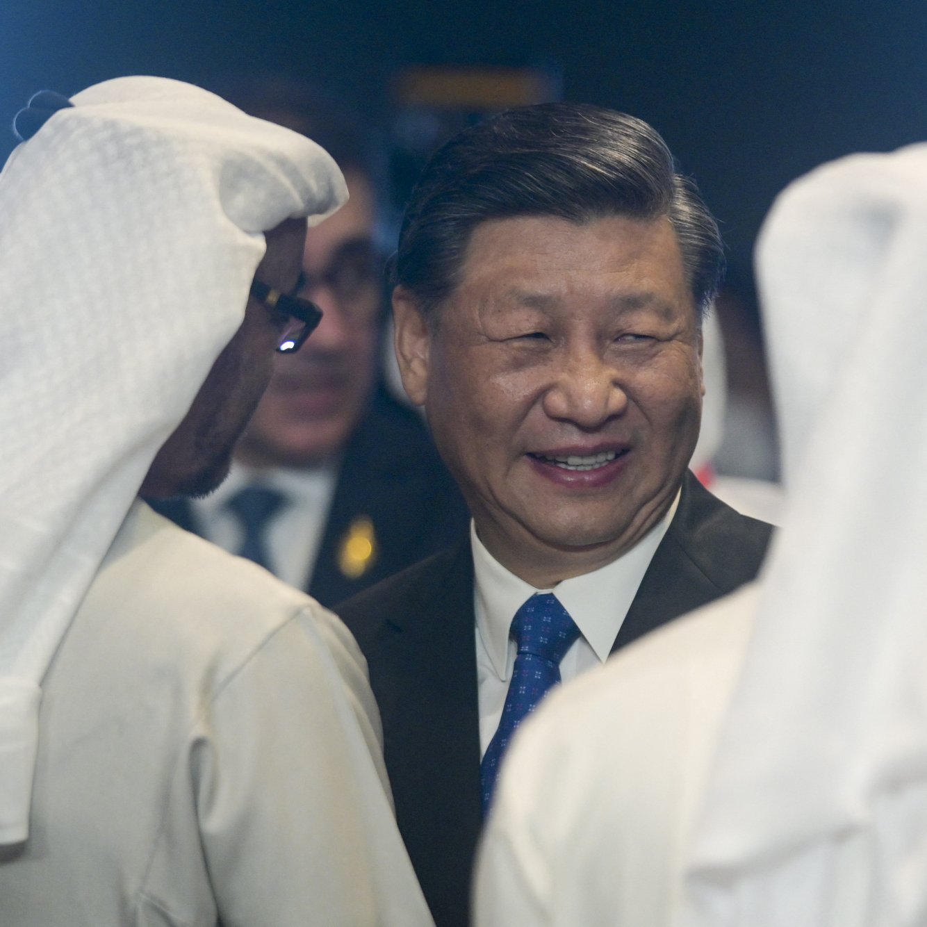 L'image montre un homme souriant, probablement un leader, en conversation avec deux autres personnes portant des turbans blancs. L'ambiance semble formelle et des éléments de l'environnement suggèrent un contexte officiel.