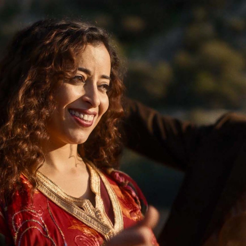 L'image montre une femme souriante avec des cheveux bouclés, portant une robe rouge ornée de motifs. Elle semble joyeuse et est entourée d'un environnement naturel. Un homme en arrière-plan, vêtu d'un costume, semble également profiter du moment. L'atmosphère générale est festive et conviviale.