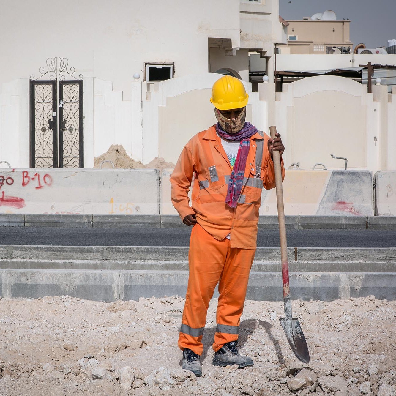 L'image montre un homme qui travaille sur un chantier de construction. Il porte un uniforme orange, ainsi qu'un casque de sécurité jaune. Il semble tenir une pelle, regardant vers le sol avec une posture réfléchie. En arrière-plan, on peut voir des murs de construction et des graffitis sur des surfaces environnantes. Le site semble en cours de développement, avec des matériaux de construction dispersés au sol.