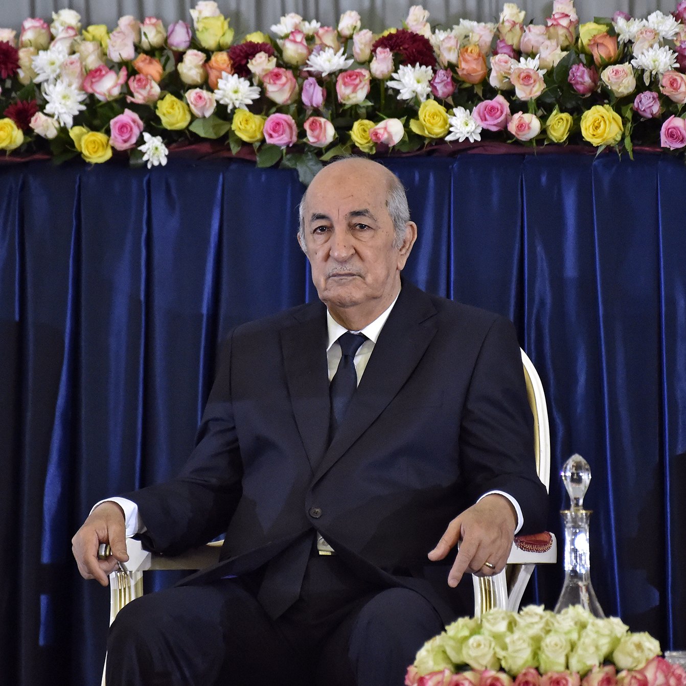 L'image montre un homme assis sur une chaise, portant un costume sombre et une cravate. Il a l'air sérieux et attentif. À l'arrière-plan, il y a une décoration florale composée de roses et d'autres fleurs qui ajoutent de la couleur à la scène. La disposition des fleurs est soignée, suggérant un cadre formel ou officiel.