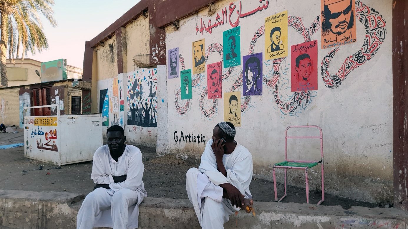 L'image montre deux hommes assis à l'extérieur, probablement dans une rue d'une ville. Ils portent des vêtements traditionnels, en particulier des robes longues. En arrière-plan, il y a un mur décoré de peintures murales colorées représentant des portraits. On peut également voir des motifs et des inscriptions en arabe. L'ambiance semble calme, avec des palmiers visibles en haut de l'image, suggérant une atmosphère chaleureuse.