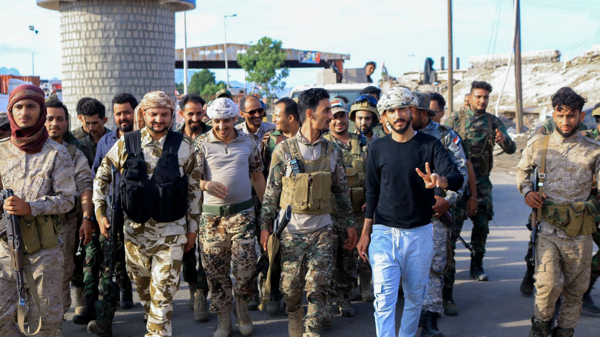 L'image montre un groupe de soldats marchant ensemble dans une zone urbaine. Parmi eux, on peut voir des militaires en uniformes camouflés et d'autres en tenues militaires variées. Certains portent des gilets tactiques et des casques. On remarque une atmosphère de camaraderie et de détermination parmi les membres du groupe, avec une personne au centre faisant un signe de paix. En arrière-plan, des infrastructures urbaines sont visibles, ce qui indique un environnement de mission militaire.