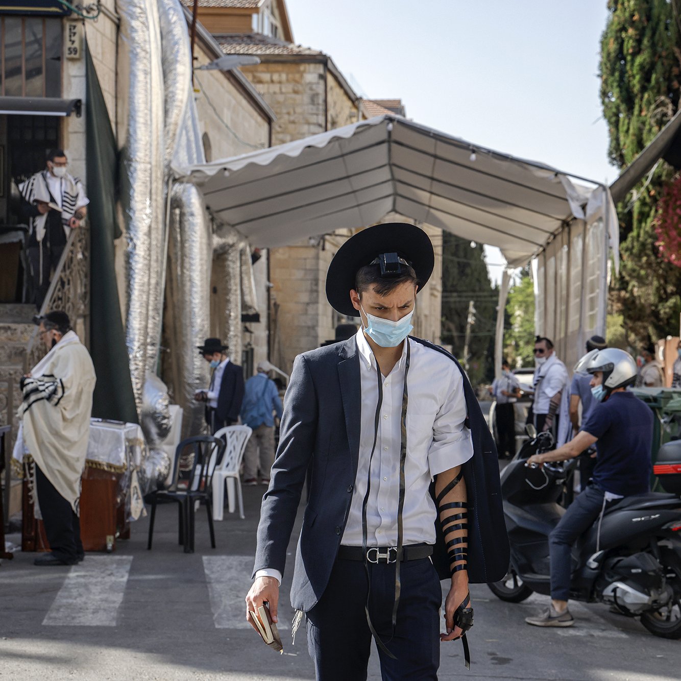 The image shows a street scene in an urban area, likely in a Jewish neighborhood. A young man wearing a mask and traditional attire, including a black hat and a suit, is walking while wearing tefillin (phylacteries) on his arm. In the background, there is a large tent set up for what appears to be a religious gathering, with several other individuals, some dressed in traditional clothing, visible. The setting is sunny, and there are various buildings, including signs and greenery, contributing to the atmosphere of the scene.