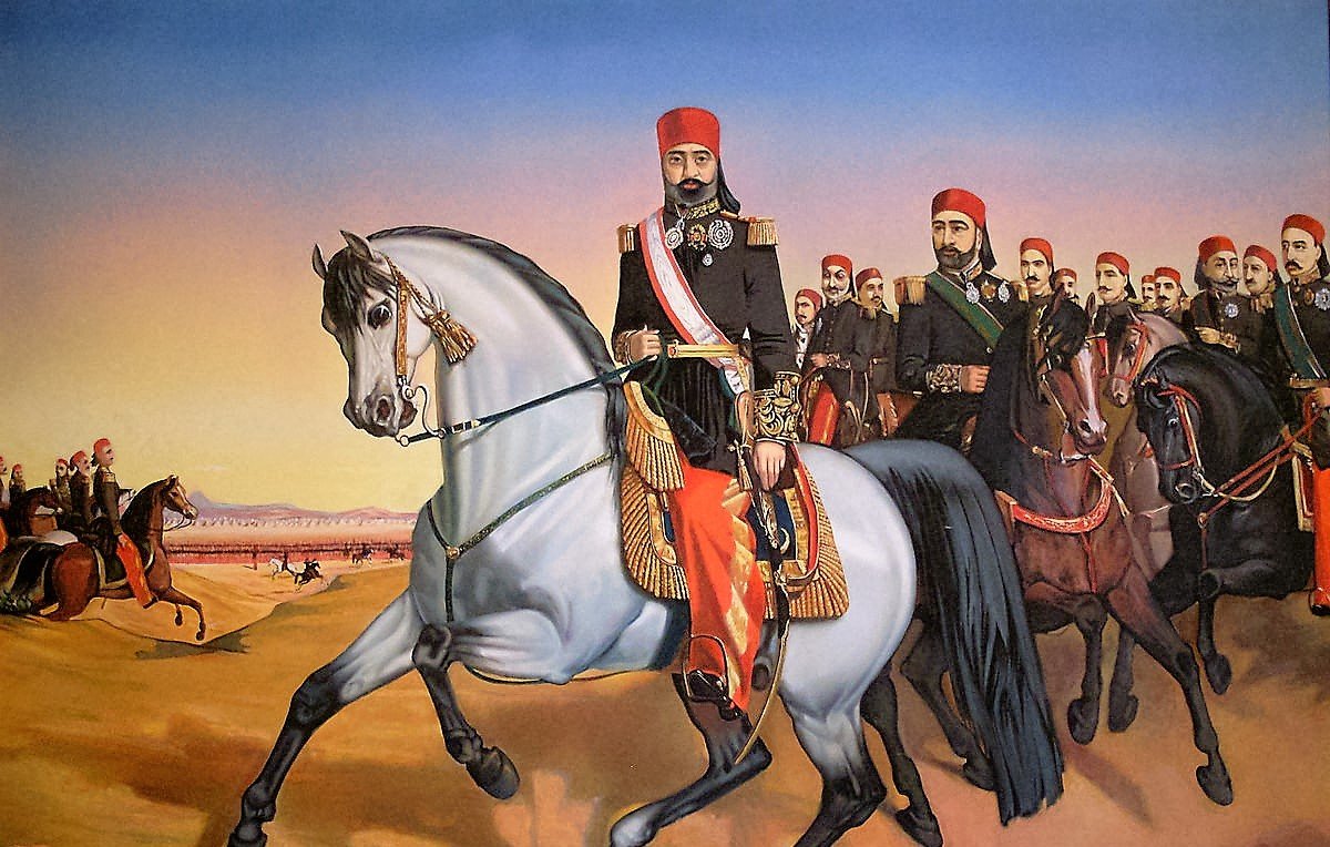 L'image représente un groupe de cavaliers en tenue traditionnelle, menés par un homme à cheval en avant-plan. Cet homme, qui semble être une figure d'autorité, est vêtu d'un uniforme orné et porte un chapeau distinctif. Il est monté sur un cheval gris, tandis qu'un groupe d'hommes sur des chevaux bruns et noirs le suit derrière lui. Le paysage est désertique, avec des montagnes en arrière-plan et un ciel aux teintes chaudes, suggérant le lever ou le coucher du soleil. Cette scène dégage une impression de mouvement et de puissance.