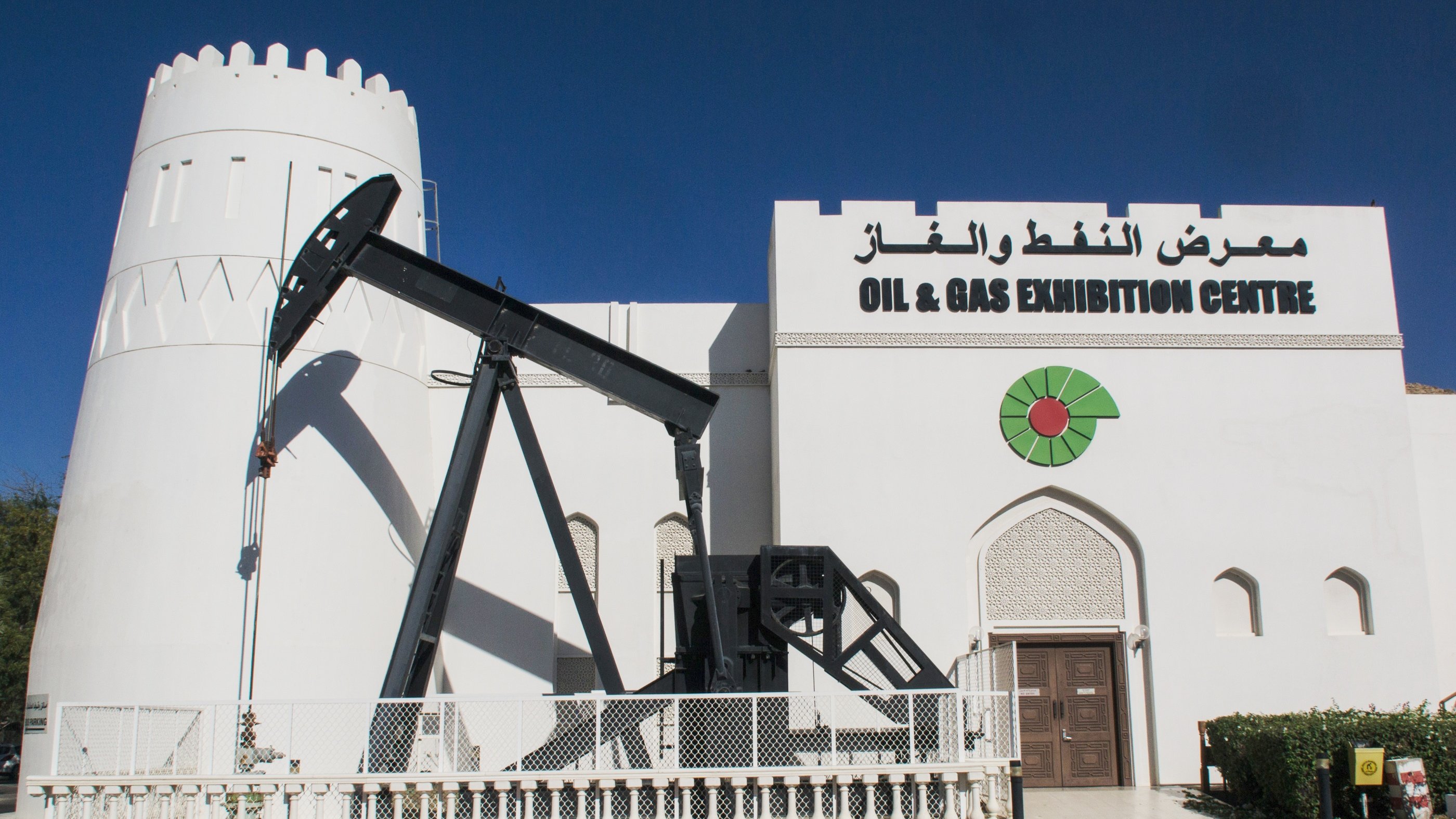 الصورة تظهر مبنى يبدو كمعرض للنفط والغاز، حيث يتضمن تصميمه معالم معمارية مميزة مثل جدران بيضاء وأبراج. أمام المبنى يوجد جهاز استخراج النفط المعروف بـ "رافعة النفط" الذي يُستخدم في عمليات الحفر والتنقيب عن النفط. كما يُلاحظ في التصميم وجود شعار معبر يحتوي على رمز يشير إلى الطاقة، مع كلمات تشير إلى أهمية المعرض في مجال النفط والغاز. السماء في الصورة واضحة والجو يبدو مشمسًا.