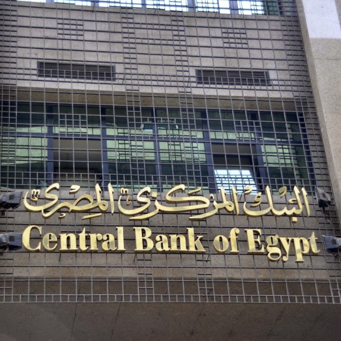 L'image montre la façade d'un bâtiment moderne, probablement celle de la Banque centrale d'Égypte. On peut voir le nom de l'institution écrit en arabe et en anglais, avec un design sobre et élégant. La structure est faite de granite et présente des grilles métalliques sur les fenêtres, ce qui donne une impression de sécurité et de solidité.
