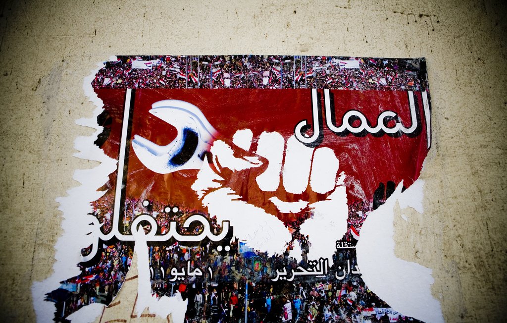 L'image montre une affiche partiellement déchirée qui présente un poing levé, symbole de révolte ou de solidarité. En arrière-plan, on peut apercevoir une grande foule, probablement lors d'une manifestation. Le fond est rouge, et il y a des inscriptions en arabe et en d'autres langues qui semblent exprimer des messages politiques ou sociaux. Le tout évoque un fort sentiment de lutte collective et de volonté de changement.