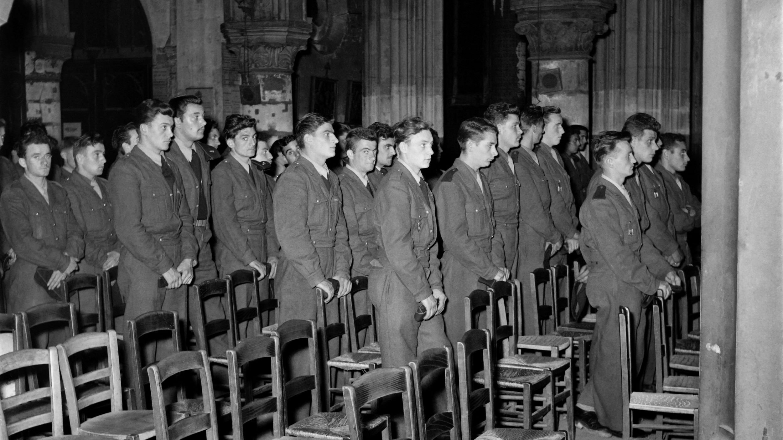 L'image montre un groupe de jeunes hommes en uniforme, probablement des soldats, se tenant debout en rang dans une église ou un bâtiment religieux. Ils semblent attentifs et concentrés, se tenant droit, dans un environnement décoré d'éléments architecturaux gothiques. Des chaises en bois sont visibles devant eux, et l'éclairage met en valeur l'atmosphère solennelle du lieu.