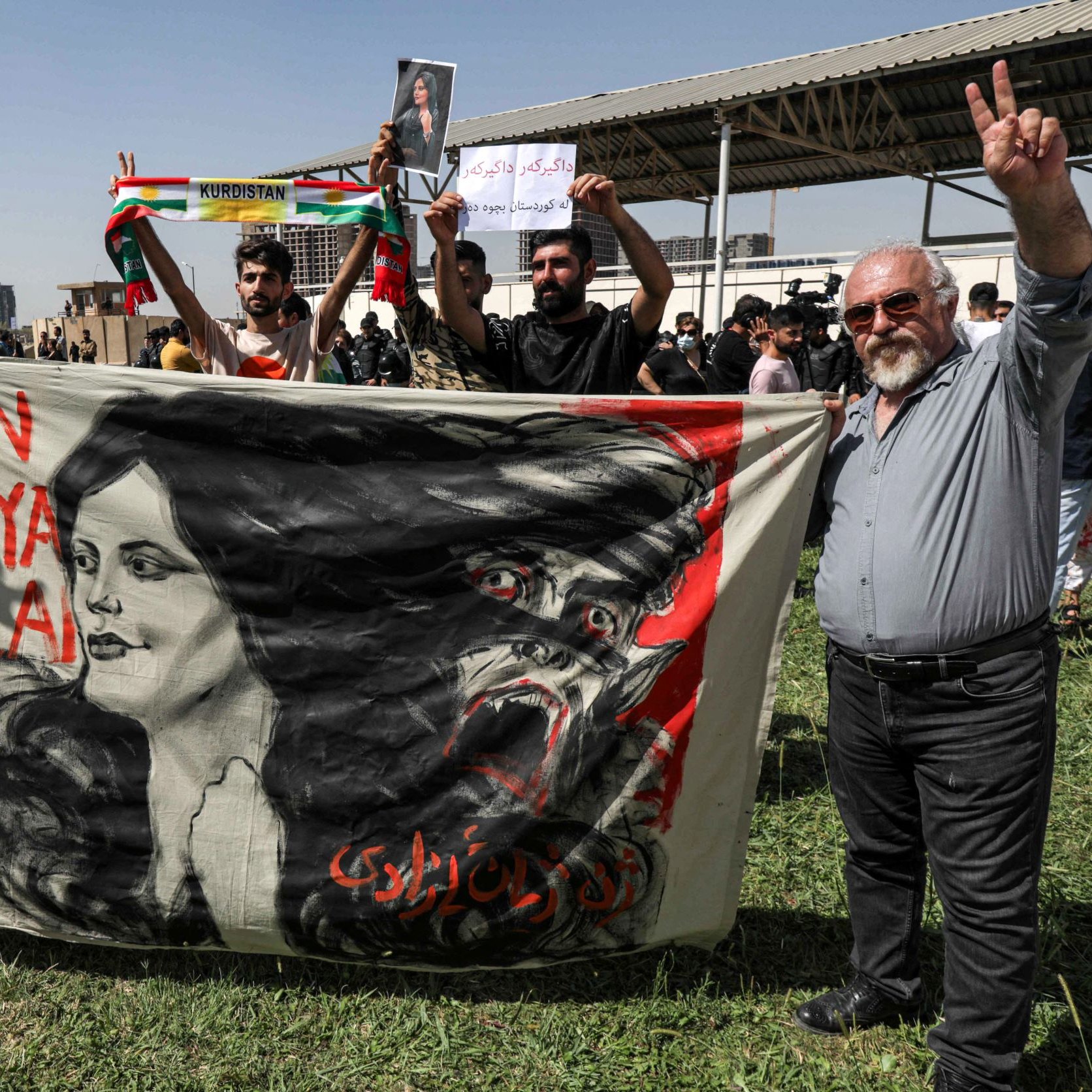 در این تصویر، افراد مختلفی در یک تجمع یا تظاهرات حضور دارند. دو نفر در جلو در حال نمایش یک بنر بزرگ هستند که تصویر زنی با نام "زن، زندگی، آزادی" و یک سر حیوان را نشان می‌دهد. در پس‌زمینه، عده‌ای دیگر در حال برگزاری تظاهرات هستند و پلاکاردهایی در دست دارند. فضای تصویر نشان‌دهنده همبستگی و جنبش‌های اجتماعی است. افراد در حال نشان دادن علامت پیروزی هستند و احساسات قوی را ابراز می‌کنند.