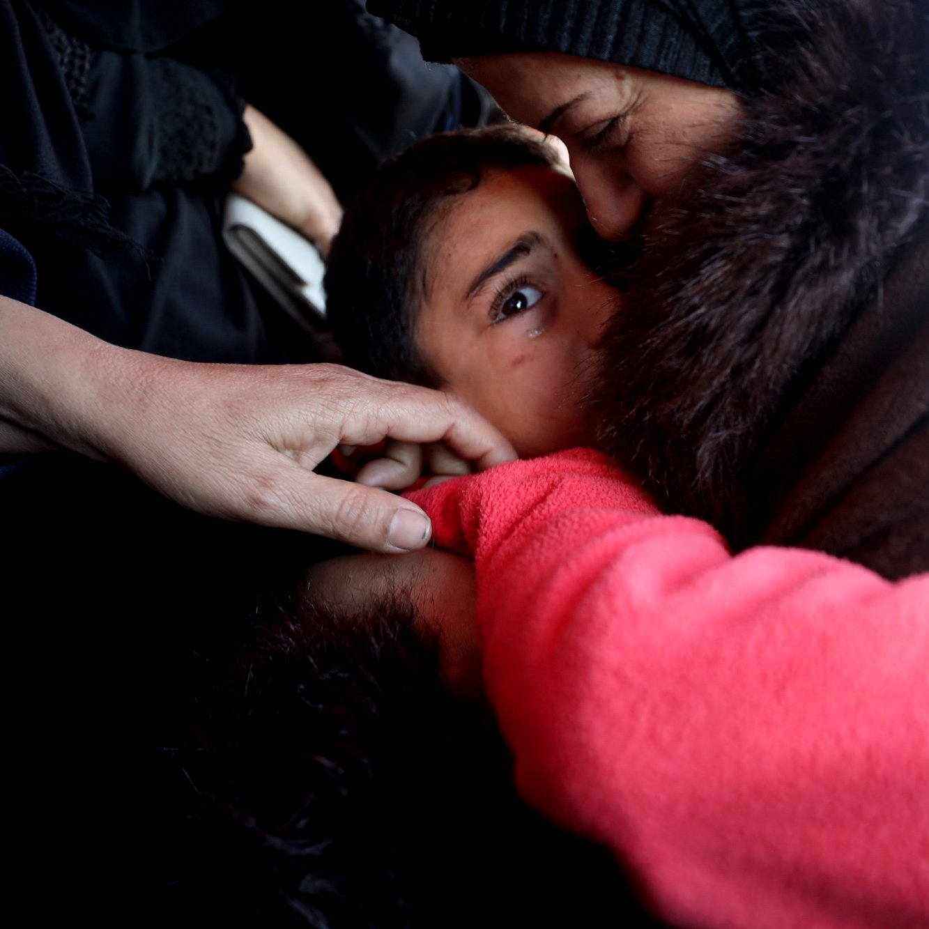L'immagine mostra un momento intimo e toccante tra un adulto e un bambino. Il bambino, con uno sguardo curioso e attento, è avvolto in un abbraccio mentre l'adulto, probabilmente una madre o una figura di riferimento, lo bacia affettuosamente. Le mani di un'altra persona si intravedono, creando una sensazione di protezione e vicinanza. L'atmosfera trasmette emozione e calore umano, evidenziando il legame profondo tra le persone. La scena è caratterizzata da toni scuri e un'illuminazione morbida, che contribuiscono a enfatizzare la tenerezza del momento.