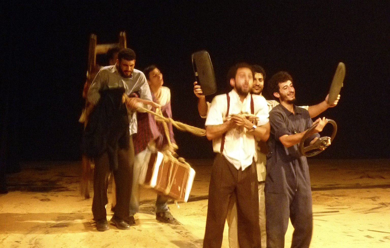 L'image montre un groupe de personnes sur une scène de théâtre, en train de créer une sorte de performance. Ils semblent emporter différents objets, comme des chaises et une valise, en formant une sorte de procession. La scène est éclairée de manière à mettre en évidence leur mouvement, tandis que le fond est sombre, ce qui attire l'attention sur les acteurs. L'ensemble donne une impression dynamique et dramatique, évoquant une histoire ou une interaction entre les personnages.