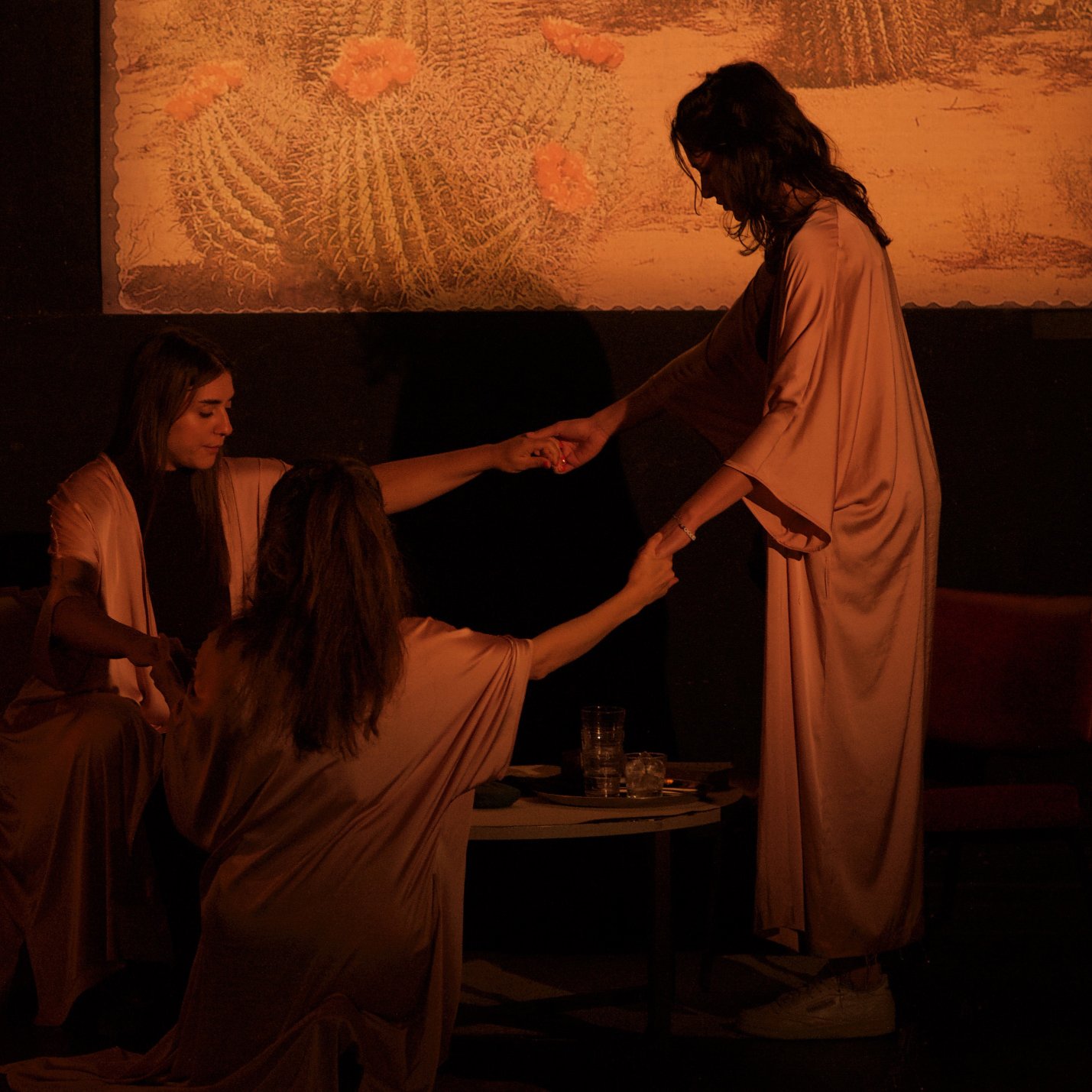 L'image présente une scène théâtrale ou artistique, où trois femmes en vêtements fluides de couleur beige se tiennent autour d'une table basse. Elles semblent engagées dans une interaction attentive, tenant les mains les unes des autres. La lumière est tamisée, créant une atmosphère intime et mystérieuse. En arrière-plan, il y a un tableau représentant des cactus dans un paysage désertique, ajoutant une touche de nature à la scène. Les expressions des femmes ne sont pas clairement visibles, contribuant à l'ambiance intrigante de l'image.