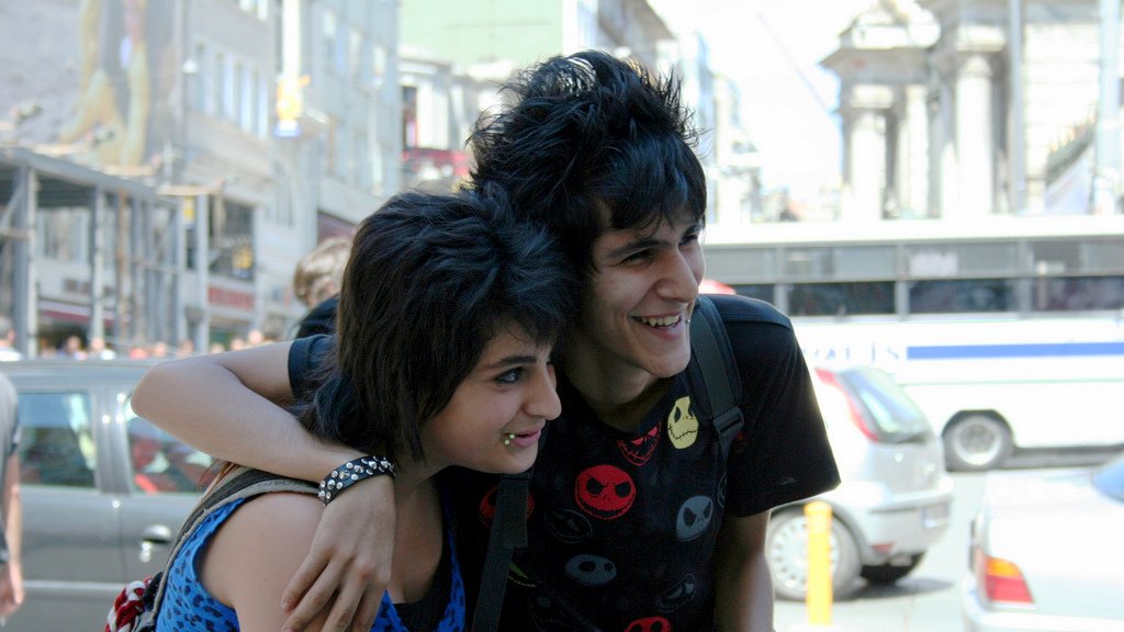 L'image montre deux jeunes personnes souriantes, posant côte à côte dans une rue animée. L'un des jeunes porte un t-shirt noir à motifs colorés, tandis que l'autre a un haut à motifs bleus. Ils semblent heureux et proches, et l'ambiance est vivante, avec des voitures et des piétons en arrière-plan. Les bâtiments en arrière-plan ajoutent à l'environnement urbain.