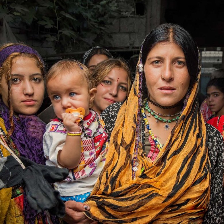 L'image montre un groupe de femmes et d'enfants, probablement dans un environnement rural. Les femmes portent des vêtements colorés et traditionnels, avec des foulards sur la tête. L'une d'elles tient un bébé dans ses bras. Les expressions sur leurs visages sont variées, allant de la curiosité à la détermination. On peut aussi voir des éléments culturels dans leur tenue, renforçant l'idée d'une riche tradition. L'arrière-plan semble un peu flou, suggérant un cadre rural ou communautaire.