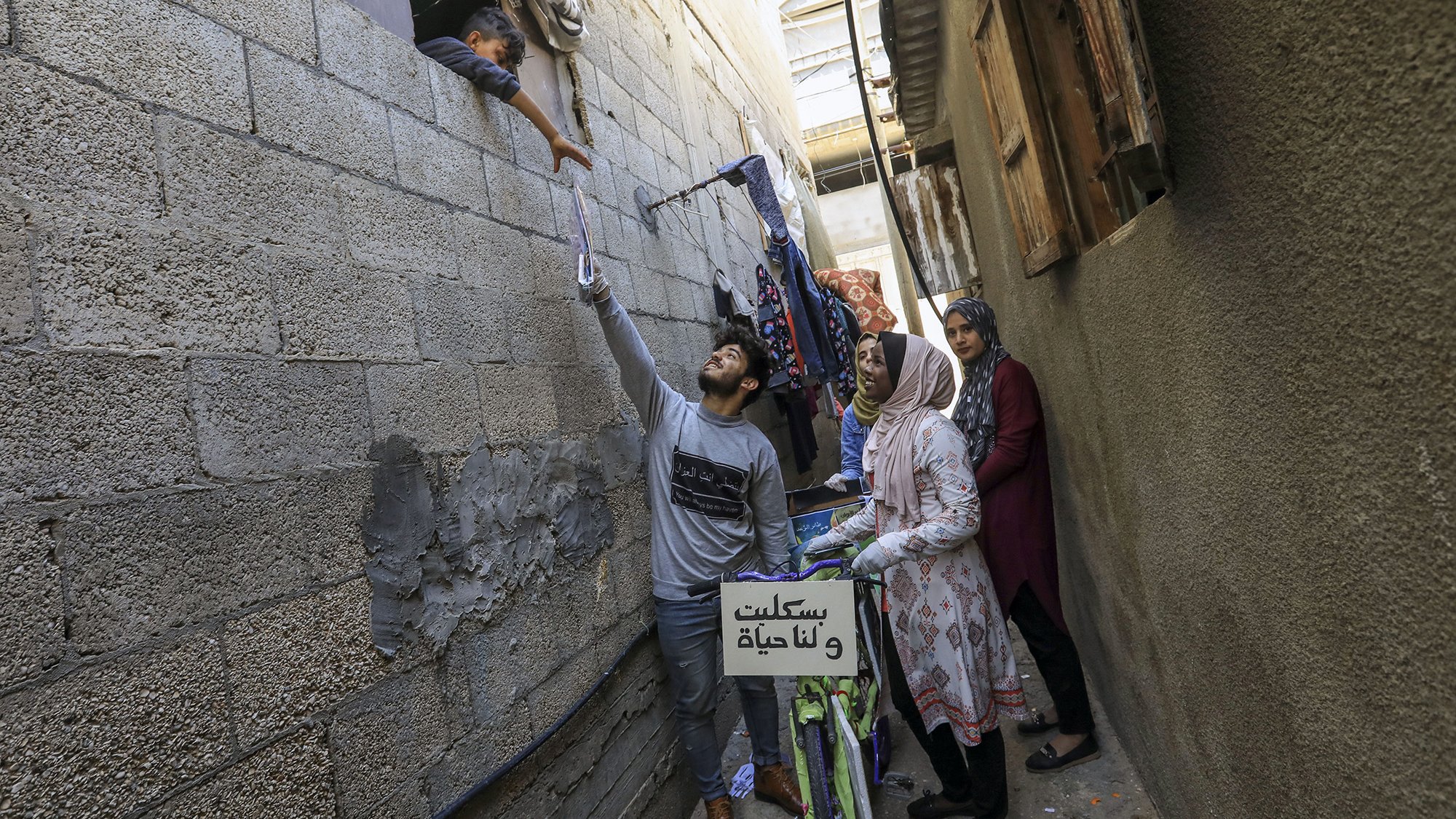 L'image montre une scène dans une ruelle étroite, probablement dans un quartier urbain. On y voit un jeune homme qui tend la main vers une fenêtre, où un enfant semble interagir avec lui. À l'arrière-plan, une femme, probablement la mère de l'enfant, observe la scène. Une autre femme se tient à côté, regardant également. Des vêtements sont suspendus à l'extérieur, ajoutant une touche de vie quotidienne à l'image. Une bicyclette est visible, avec un panneau sur le guidon. L'atmosphère semble chaleureuse et familiale, malgré le contexte urbain.
