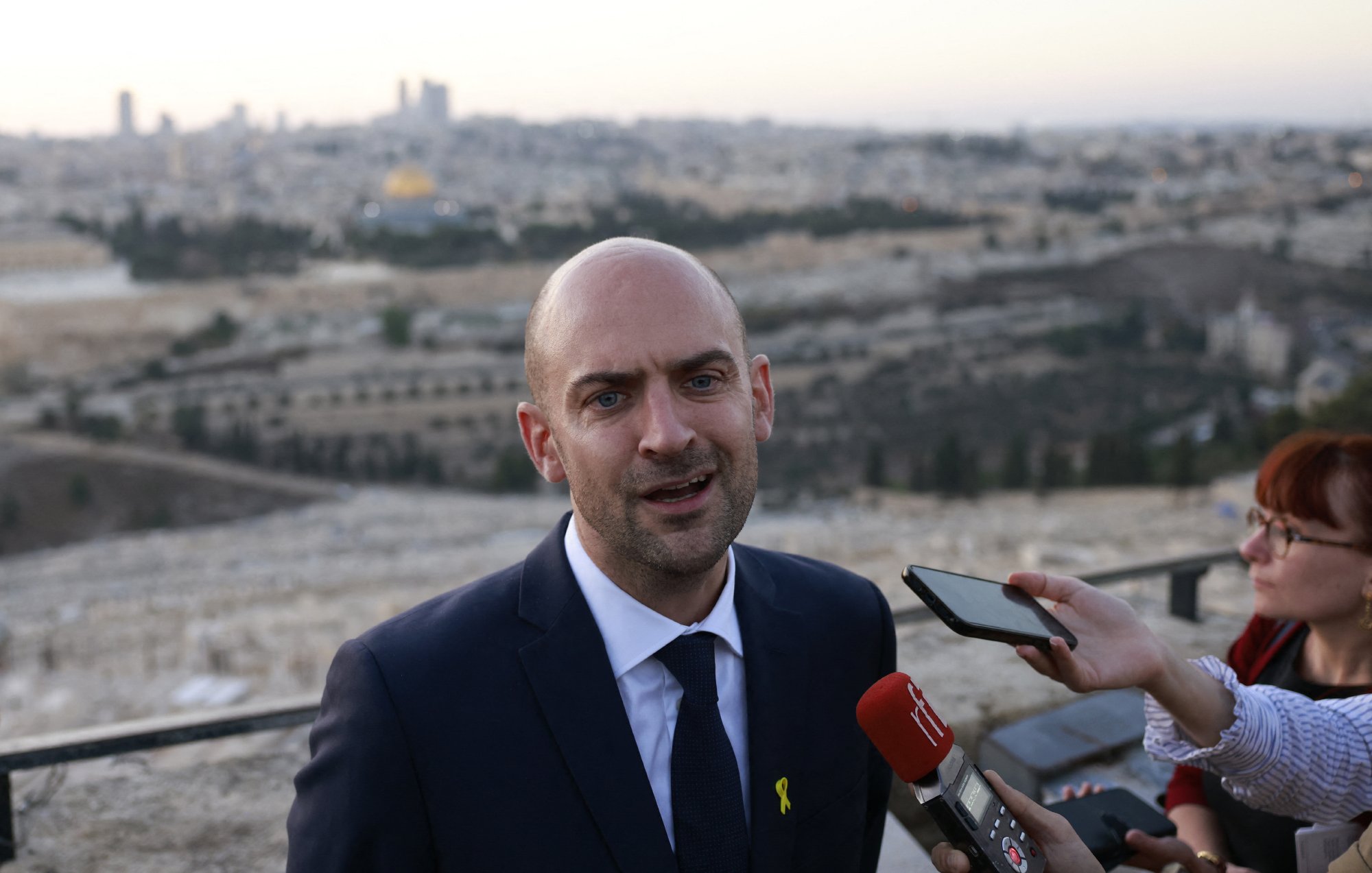 L'image montre un homme en costume, probablement un porte-parole ou un responsable politique, s'exprimant devant un groupe de journalistes. Il se tient à l'extérieur avec une vue panoramique de la ville en arrière-plan, qui semble être Jérusalem, avec des bâtiments et un paysage urbain. Le ciel est clair, et la lumière du crépuscule donne une atmosphère calme à la scène. Plusieurs microphones et téléphones montrent l'intérêt médiatique pour ses déclarations.