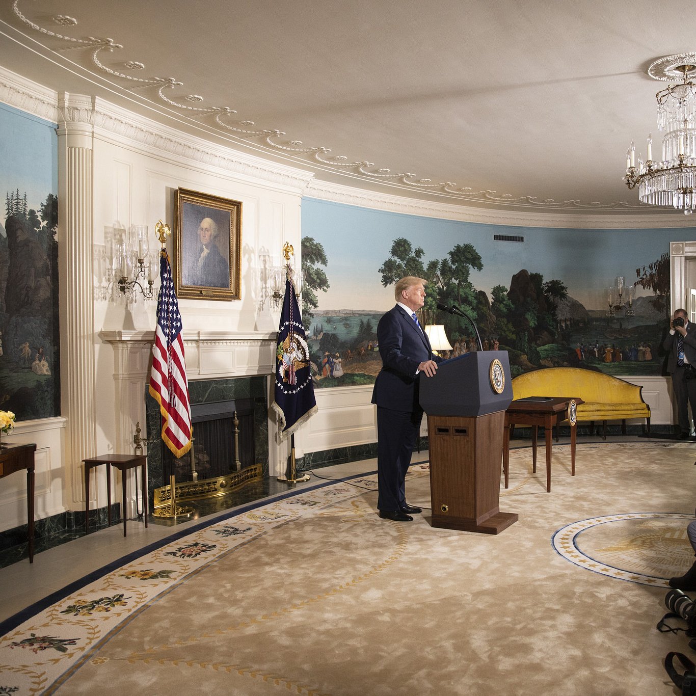در این تصویر، دونالد ترامپ در حال سخنرانی است و در پس‌زمینه، دیواری با نقاشی‌های منظره و یک پرچم ایالات متحده و یک پرچم دیگر دیده می‌شود. او در کنار یک تریبون ایستاده و دوربین‌های خبرنگاران و عکاسان در جلو او قرار دارند. دکوراسیون اتاق زیبا و مجلل است، با یک فرش بزرگ و مجسمه‌هایی در اطراف. نور طبیعی از پنجره‌ها به داخل اتاق می‌تابد.